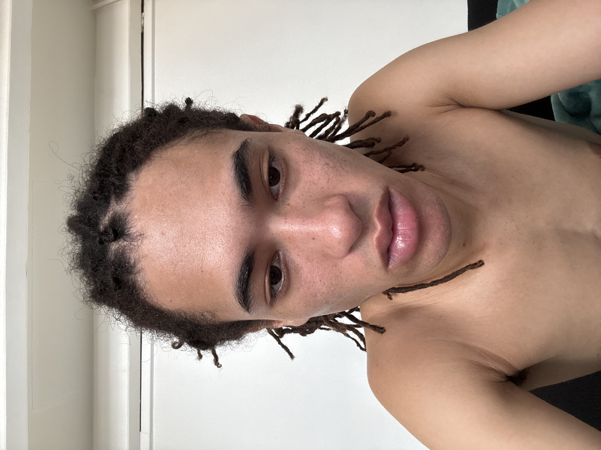 A close-up, head-and-shoulders portrait shows a young person with dark, curly hair styled into dreadlocks. They are looking down and slightly to the right, with their mouth slightly open. The background is a plain, light-colored wall with a white trim at the top. The person is shirtless, and their skin appears to have some light freckling and blemishes. They are likely indoors, in a domestic setting. There is no indication of the image being taken in Paris, France.