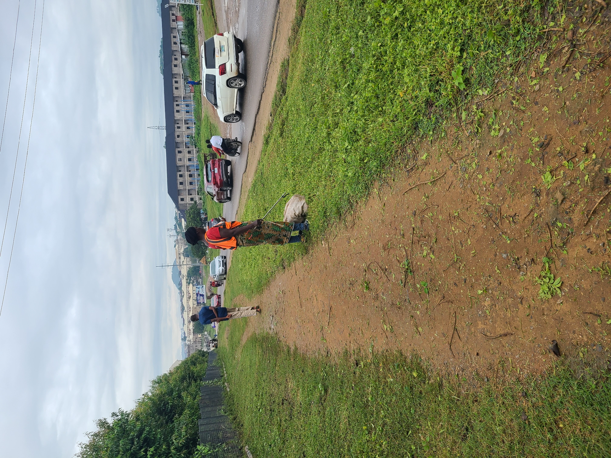 The image captures an outdoor scene in Abuja, Nigeria, featuring a dirt path adjacent to a grassy area and a road. On the path, a person wearing a bright orange vest and a patterned skirt is visible, holding a tool and a sack, suggesting they are involved in clearing debris or maintaining the area. Another individual in a blue shirt and light-colored trousers walks away from the camera on the same path, heading towards the road. The road itself has several vehicles, including a white SUV, a red car, and a motorcycle with two passengers. In the background, there are buildings, power lines, and lush green trees, indicating an urban or semi-urban environment with considerable vegetation. The sky is overcast with clouds. The overall scene depicts a moderately busy day with ongoing activities related to road and ground maintenance in a developing urban setting.