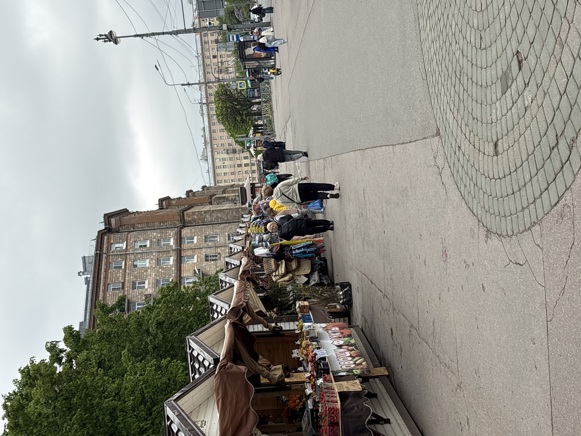 The image depicts a street scene in Saint Petersburg, Russia. Several market stalls line the right side of a wide sidewalk. The stalls appear to be selling fresh produce, including fruits and vegetables. People are browsing the stalls, possibly shopping. In the background, there is a large building. The street is paved with gray pavement and a circular brickwork. The weather appears to be overcast.