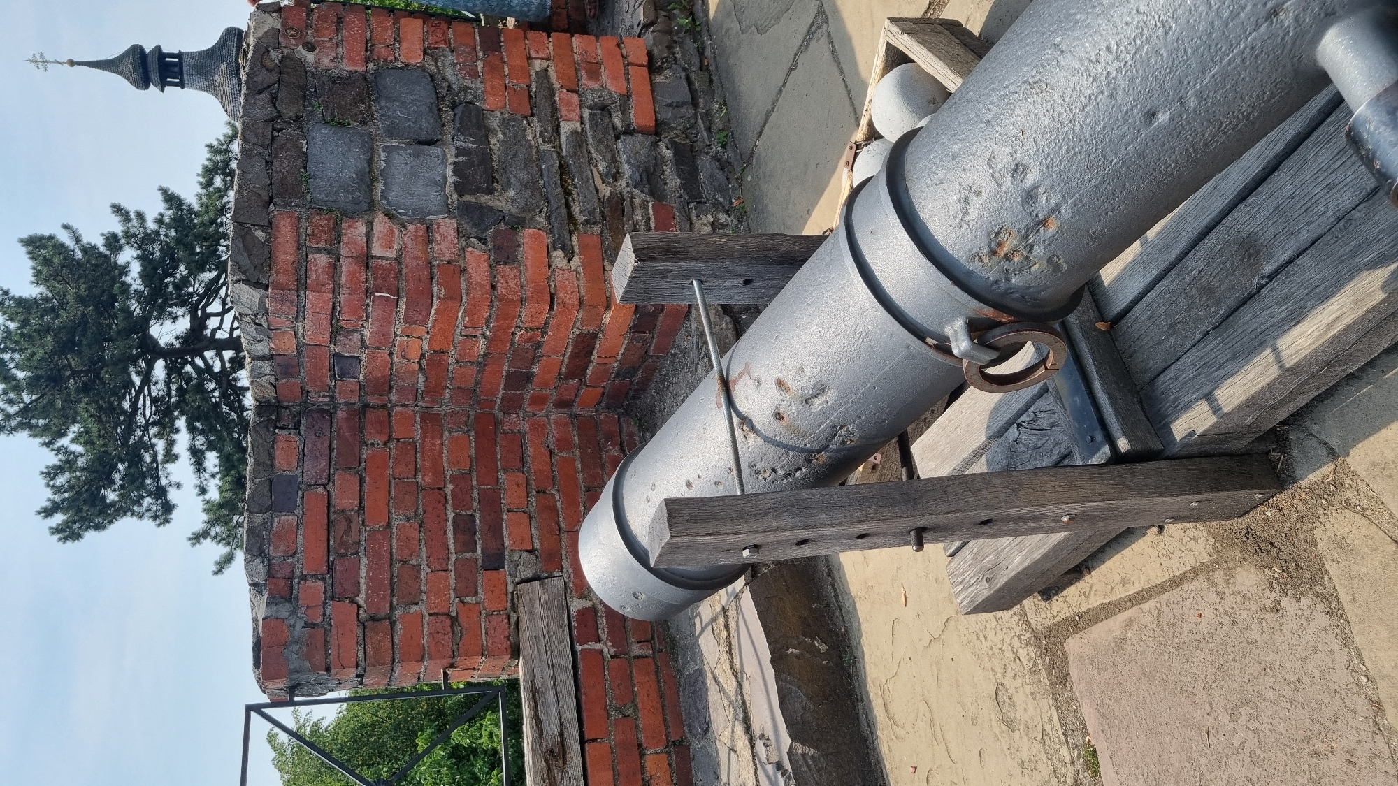 The image shows a close-up view of an old cannon positioned on a wooden carriage. The cannon is made of dark metal and is resting on a stone surface. Behind the cannon is a red brick wall and some greenery. In the background, a church spire with a cross can be seen. The scene appears to be outdoors on a sunny day. This cannon is located in Uzhhorod Castle, Uzhhorod, Ukraine.