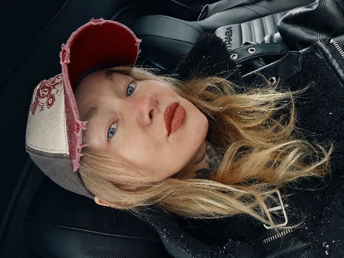 The image displays a close-up portrait of an adult female subject seated within the passenger compartment of an Abarth vehicle. The subject presents with light blonde hair, blue eyes, and reddish-orange lipstick. Tattoos, appearing as dark-inked floral or organic patterns, are visible on the left side of her neck and collarbone. She wears a distressed baseball cap featuring a dark grey rear section, an off-white front panel, and a frayed red brim with a partially visible red graphic design on the front. Her attire includes a black jacket, potentially leather or a similar material, with a textured or fur-lined collar, metallic zipper details, and a buckle on the left shoulder/chest area. Sparse white flecks, possibly snow or lint, are visible on the jacket. The vehicle's interior is dark, with a black car seat and headrest visible behind the subject, clearly displaying the white "ABARTH" branding. The subject gazes directly into the camera. No discernible external landmarks or environmental features are present to identify a specific location within the city of Saint Petersburg, Russia.