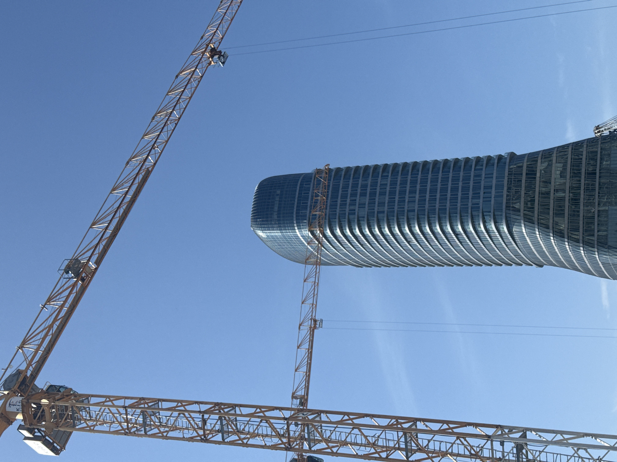 The image shows a skyscraper under construction in Belgrade, Serbia, framed by the cranes used in its construction. The sky is clear and blue. The skyscraper's architecture features curved, horizontal lines, and it is made of glass and steel. There are three cranes visible, two in the foreground and one rising along the building's facade. The crane in the foreground has an orange frame.