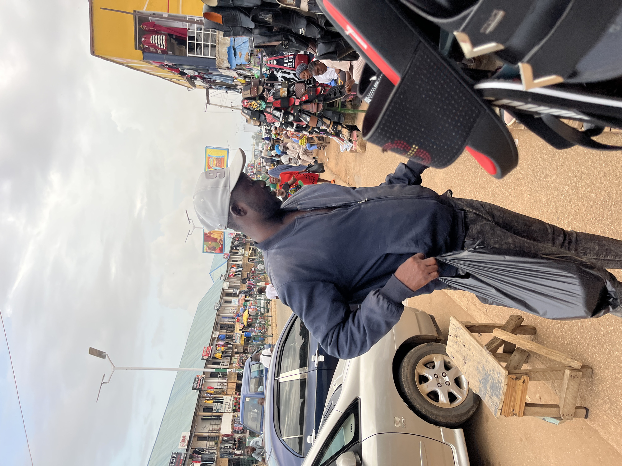 This is a medium shot of a street market in Bukuru, Nigeria. The foreground is dominated by a man wearing a gray baseball cap, a dark blue bomber jacket, and dark jeans. He is holding a black plastic bag in his right hand and appears to be looking at a display of sandals to his right. The sandals are black with red accents and are arranged on a rack.
In the background, a row of shops line the street. The shops have awnings and are filled with various goods, including clothing. Several people are visible walking along the street and interacting with vendors. Cars, including a silver sedan and a dark blue sedan, are parked on the side of the road. The ground is covered in red dirt. The sky is overcast. A street lamp is visible in the middle ground.