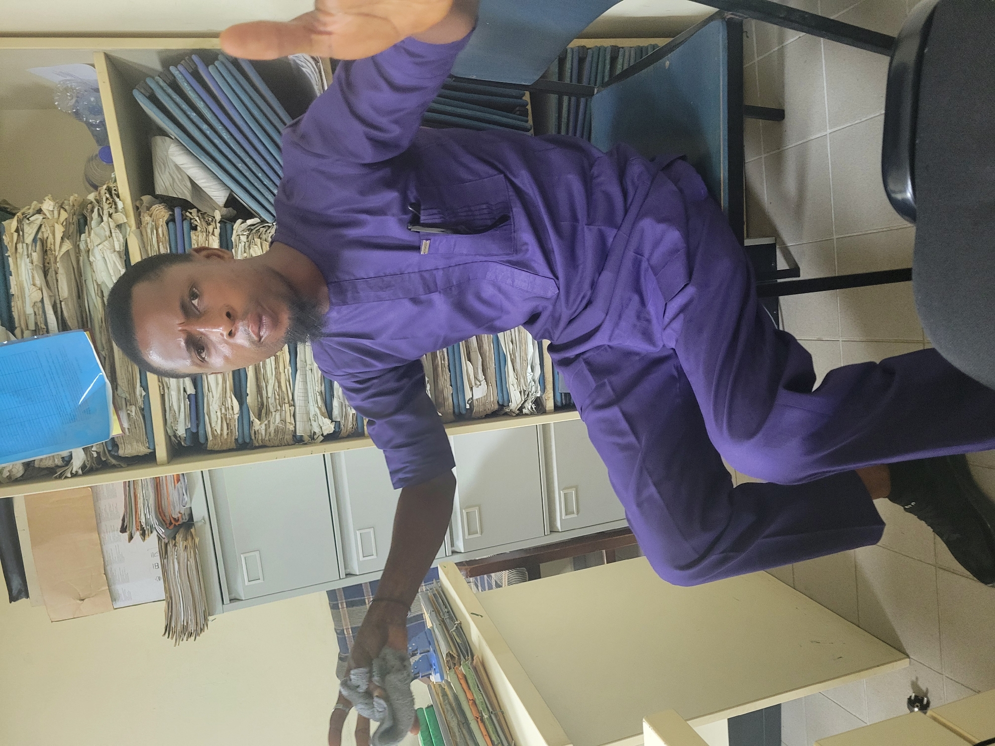 An adult male stands in an office environment in Abuja, Nigeria. He wears a purple tunic and dark shoes, is holding a gray cloth in his left hand, and extends his right hand towards the camera. Shelves filled with files and documents line the wall behind him, and several office chairs are visible. The room has beige walls and a tiled floor.