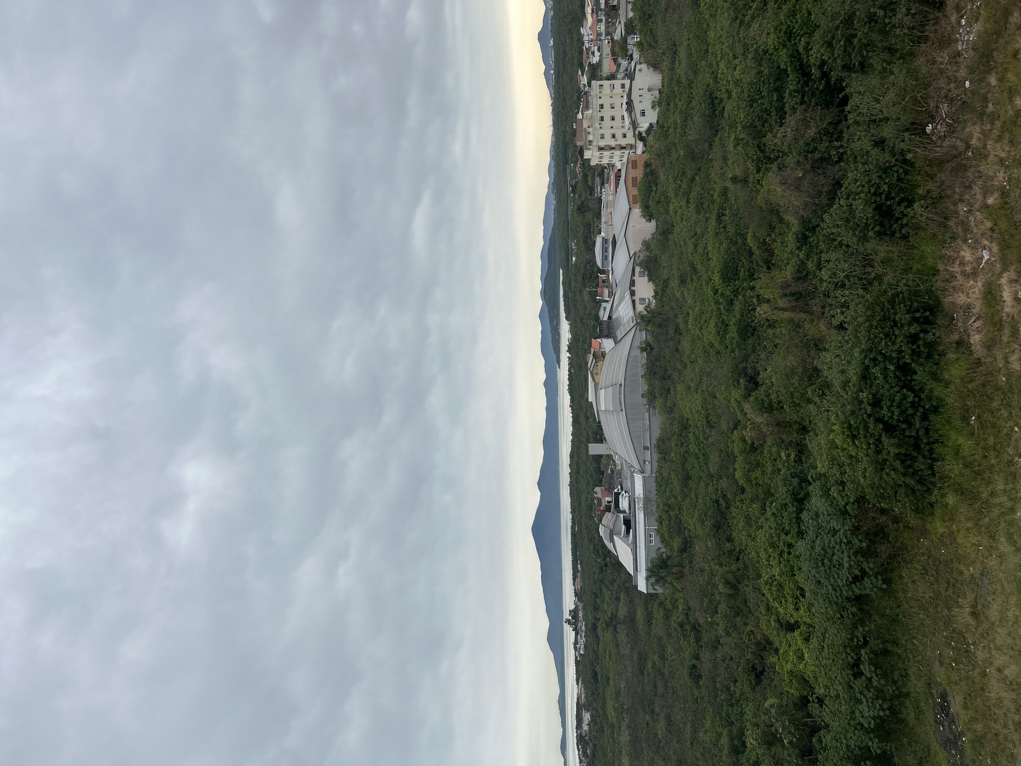 The image is a wide-angle, landscape-oriented photograph taken from an elevated position, looking down on a coastal town. The scene is divided by a horizon line: the left two-thirds are dominated by a cloudy, overcast sky, while the right third depicts a lush, green hillside dotted with buildings and overlooking a body of water and distant mountains.
The hillside is densely covered in trees and vegetation, with various structures interspersed. Closer to the viewer, there's a modern building with a curved, white roof, possibly a stadium or a large hall. Further up the hill, more buildings are visible, some appearing to be apartment complexes or hotels with multiple stories and visible windows. The architecture suggests a tropical or subtropical climate.
Below the hillside, a calm body of water stretches towards the horizon, flanked by a series of progressively larger, darker mountain ranges. The water reflects the muted light of the sky, creating a serene and slightly hazy atmosphere. The time of day appears to be late afternoon or early morning, given the soft, diffused light and the overcast conditions. The weather is clearly cloudy and somewhat dull.
The overall impression is of a developing coastal community nestled between verdant hills and the sea. There is no visible text in the image. The scene evokes a sense of tranquility and natural beauty, juxtaposed with human development.