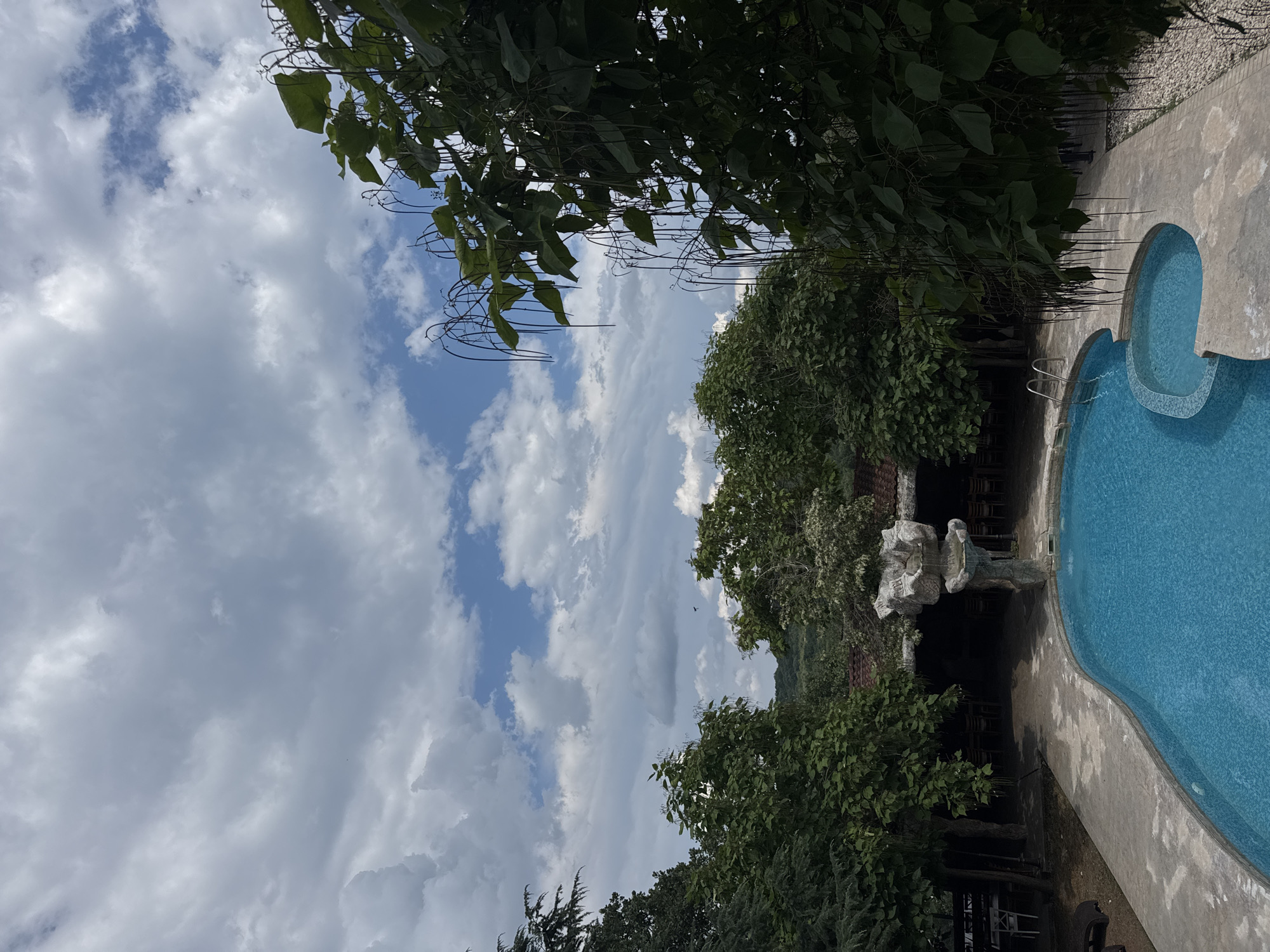 The image shows an outdoor swimming pool area with a blue pool and a smaller, integrated spa or shallow pool. The pool area is surrounded by a concrete deck and landscaping with various trees and foliage. In the background, there is a building with outdoor seating and a roof, suggesting a restaurant or recreational facility. A rock-like structure with a small waterfall feature is situated next to the pool. The sky is partly cloudy with patches of blue visible. The location is identified as being in the city of Sredets, Bulgaria. There are no people clearly visible in the immediate vicinity of the pool.