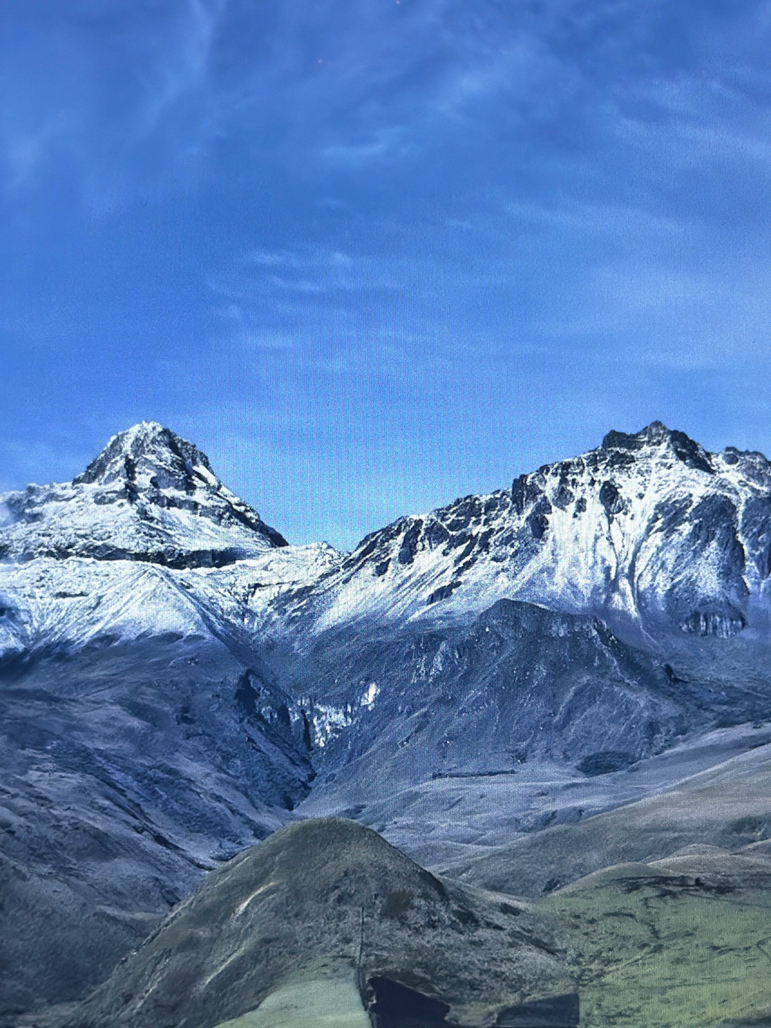 The image is a vertical, slightly grainy, medium shot of a snow-capped mountain range under a bright blue sky with wispy clouds. The mountains are rugged and appear to be volcanic in origin, with sharp peaks and steep, shadowed slopes. Snow covers the upper portions of the mountains, creating a stark contrast with the darker rock and barren earth of the lower regions. The foreground consists of rolling hills, with one prominent, darker, conical hill in the lower center. The overall lighting suggests it is daytime, with the sun high in the sky. The image does not depict any people, structures, or signs of urban life; therefore, it cannot be specifically located in Kafr Yasif, Israel.