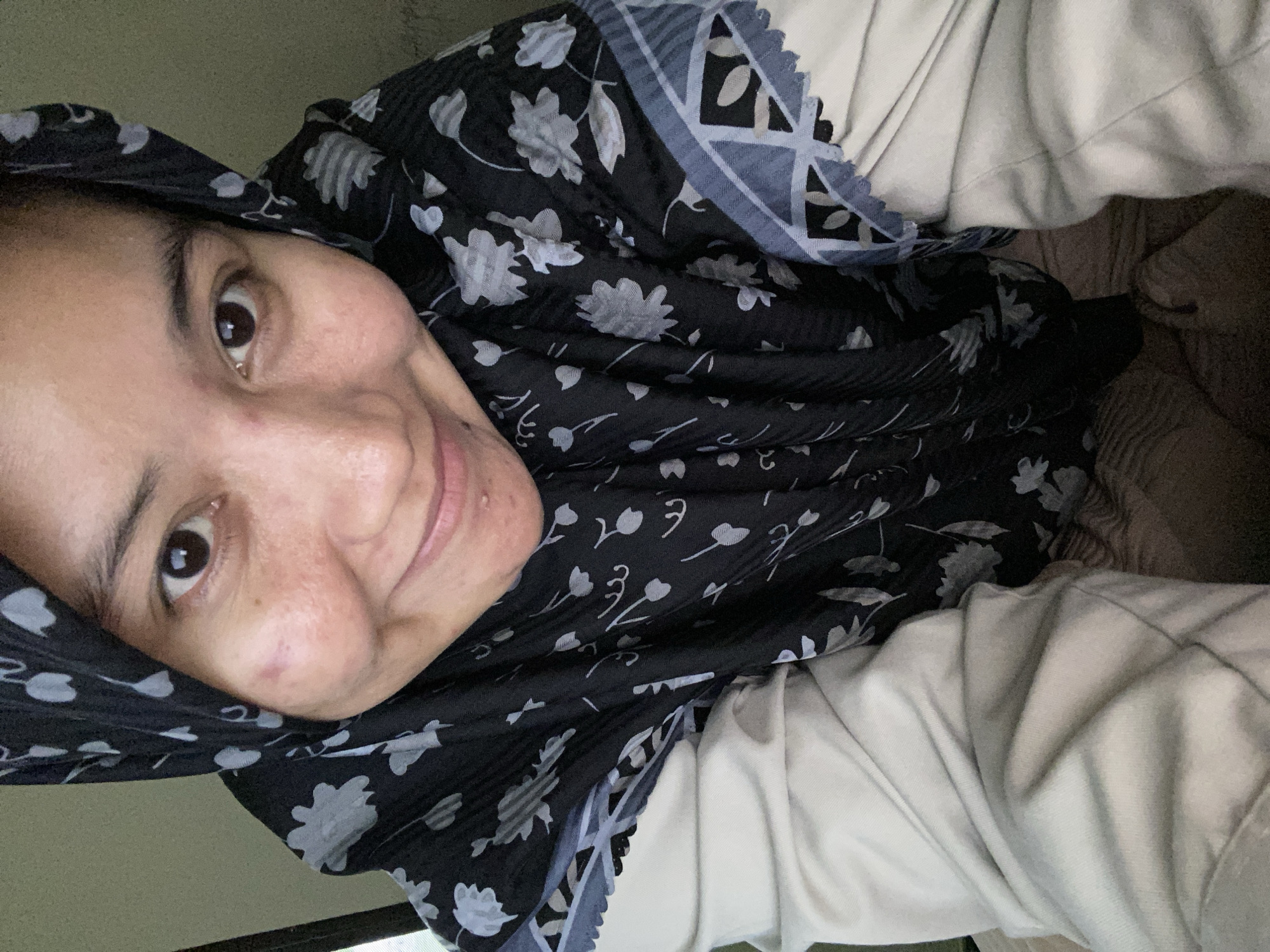 The image is a close-up, selfie-style shot of a person wearing a black hijab with a floral pattern. The person's face is visible, with one eye looking towards the camera. Their skin appears to have some texture, and there are a few small blemishes. They are smiling slightly, conveying a relaxed or content emotion. The hijab is made of a dark fabric with a repeating pattern of light-colored flowers and leaves. There is also a border with a geometric pattern along the edge of the hijab. The person is wearing a light-colored garment underneath the hijab, possibly a shirt or sweater. The background is out of focus and appears to be indoors, with soft lighting. The overall impression is personal and candid. There is no visible text in the image.
