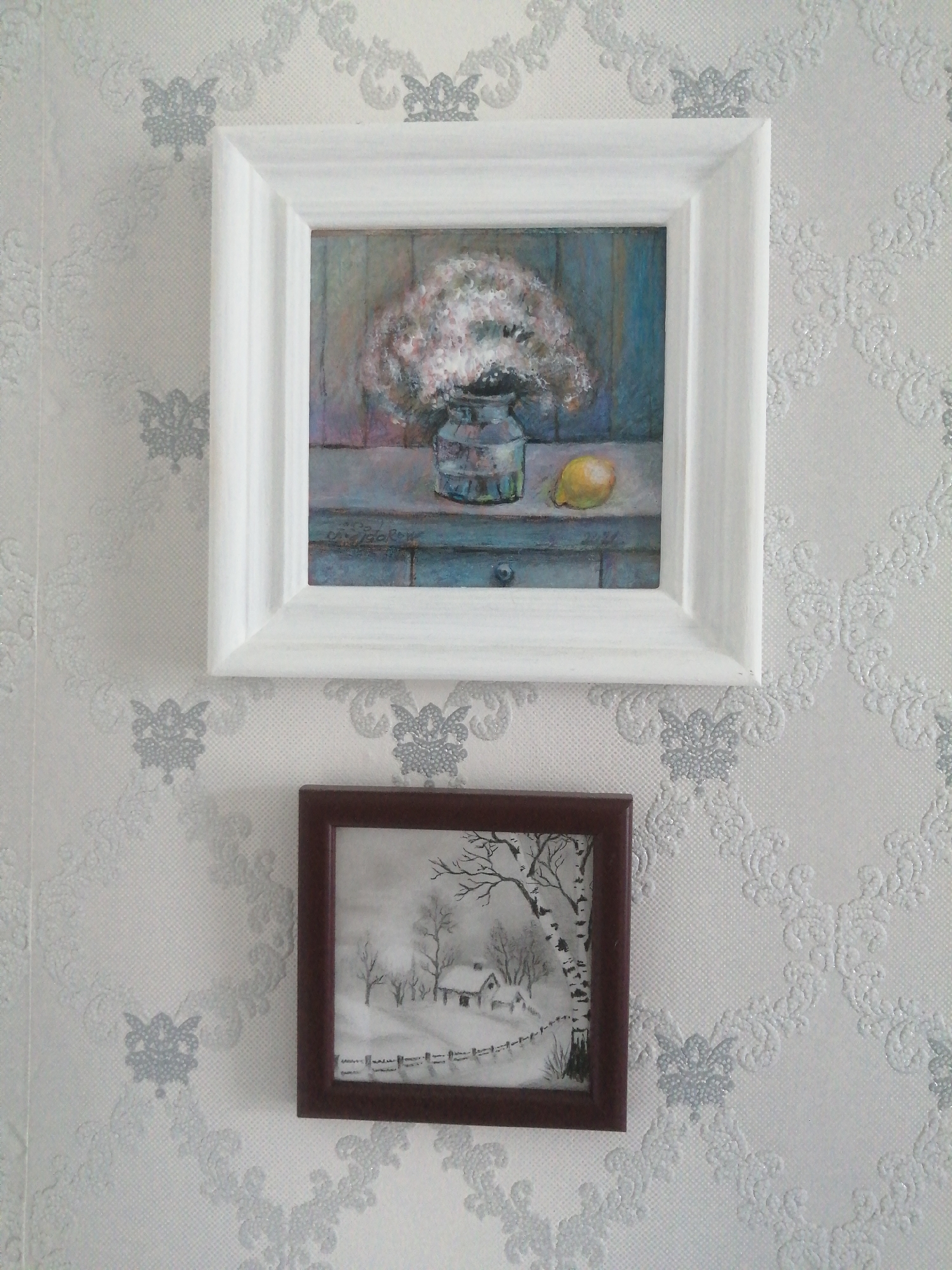 The image shows two framed pictures hanging on a wall with patterned wallpaper. The top picture is a square frame with a white border. Inside the frame is a painting of a bouquet of pink and white flowers in a blue glass vase, sitting on a blue shelf. A yellow lemon is placed to the right of the vase. The bottom picture is a smaller, square frame with a dark brown border. This frame contains a black and white drawing of a winter landscape with a small house, bare trees, and a fence. The wallpaper has a repeating pattern of light gray floral motifs. The scene appears to be indoors, possibly in a home. No specific location in Slonim, Belarus, can be identified from this image.