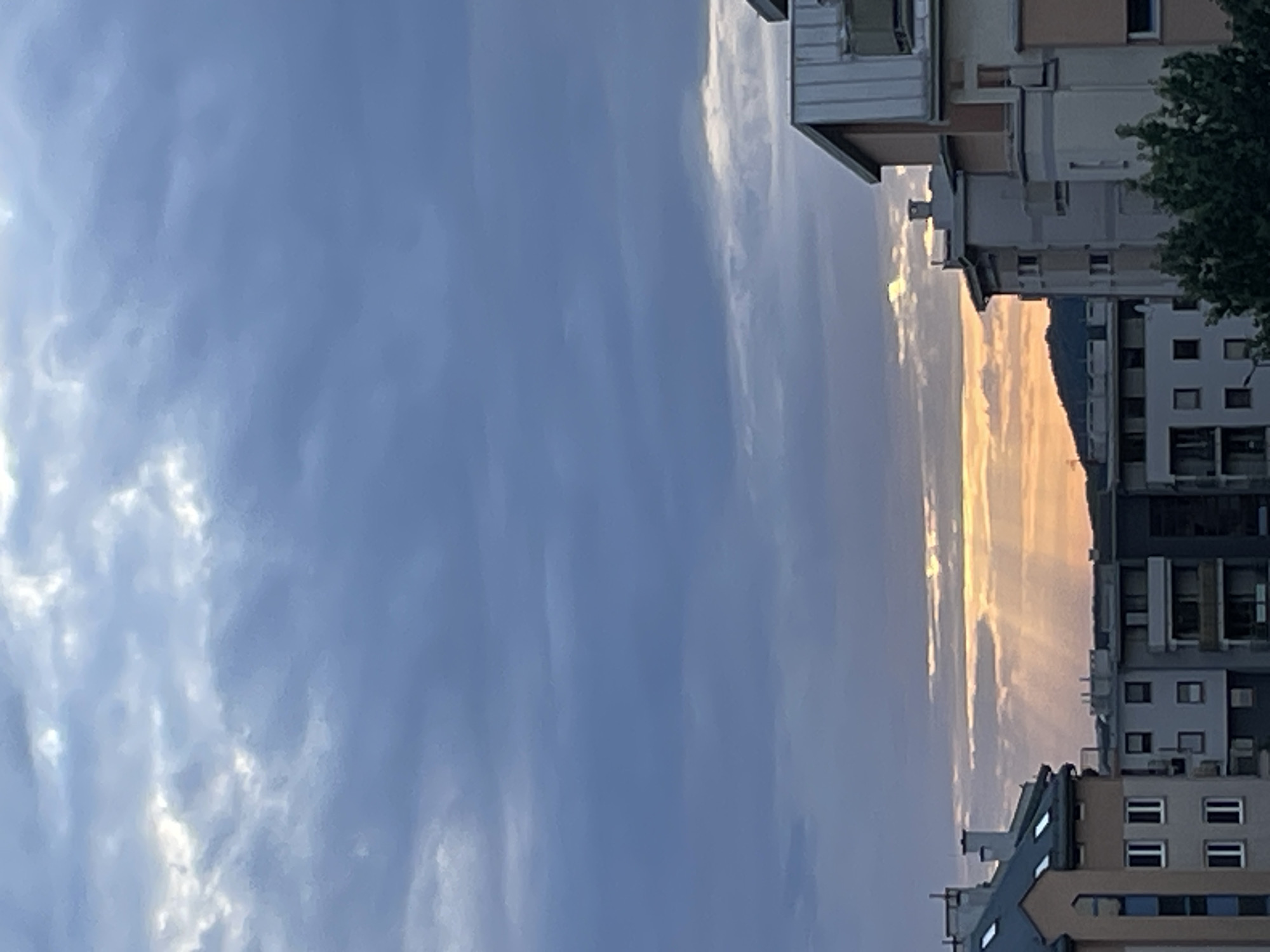 The image shows a view of buildings against a cloudy sky at sunset. The sky is dominated by shades of blue and gray clouds, with rays of golden light breaking through in the distance. The buildings are multi-story structures, some with pitched roofs and others with flat roofs. The architecture suggests an urban or suburban setting. The foreground is occupied by the tops of several buildings, with trees visible at the bottom right. The lighting suggests it is either early morning or late evening. The location is identified as Unknown, Unknown.
