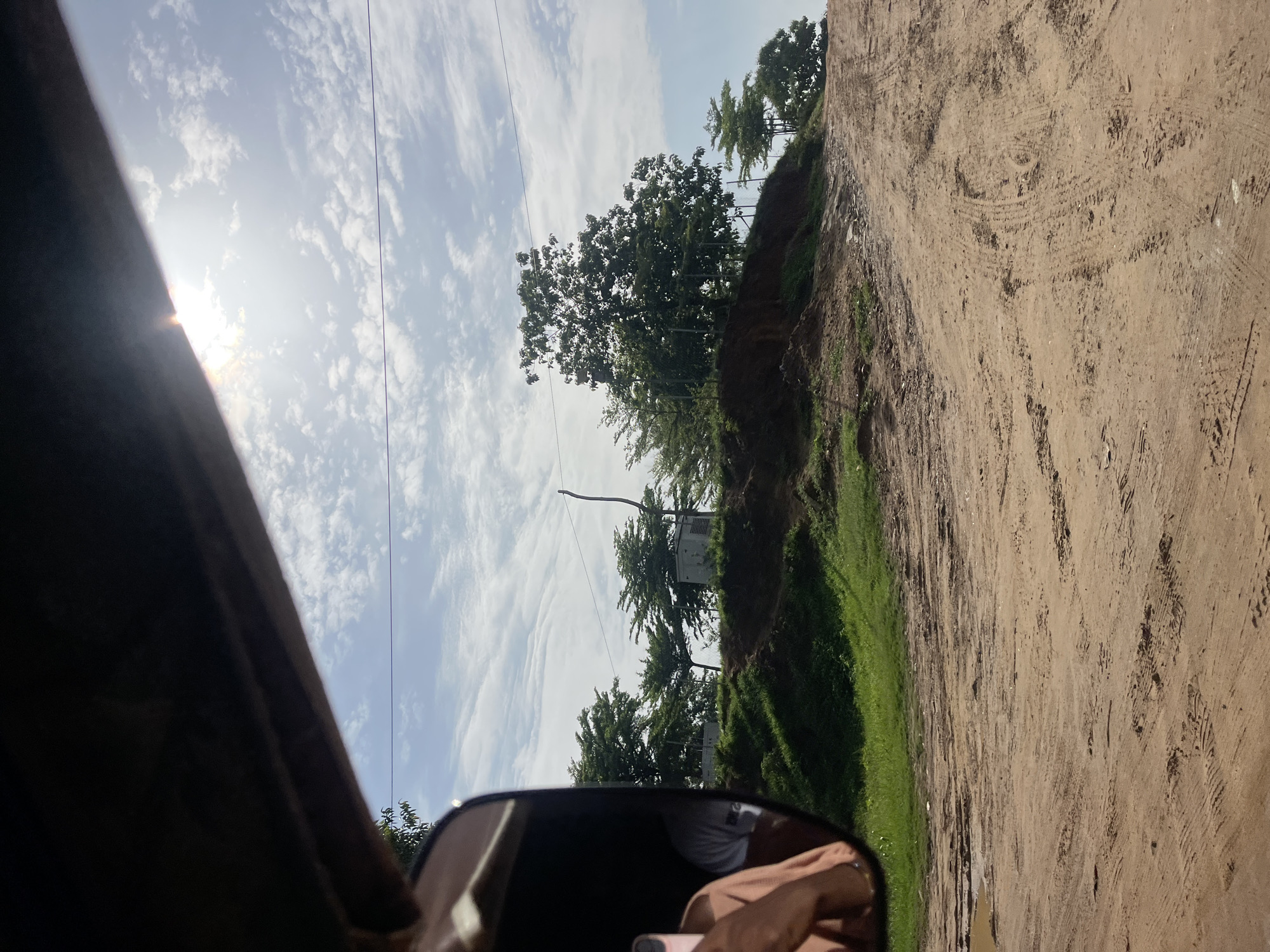 The image captures a view from a vehicle, likely a tuk-tuk, looking out at a dirt road in Karmajiji, Nigeria. The road is rutted and appears to be somewhat muddy in places, indicating recent rainfall. The left side of the frame shows a reflection in a side mirror, revealing a person's shoulder and arm, possibly the driver or passenger.
The environment is lush with green grass and several trees dotting the landscape. Some structures, possibly small buildings or utility boxes, are visible behind the trees on a raised embankment. The sky is bright with scattered white clouds and the sun is high, casting a glare. The overall impression is of a rural or semi-urban setting on a sunny day.