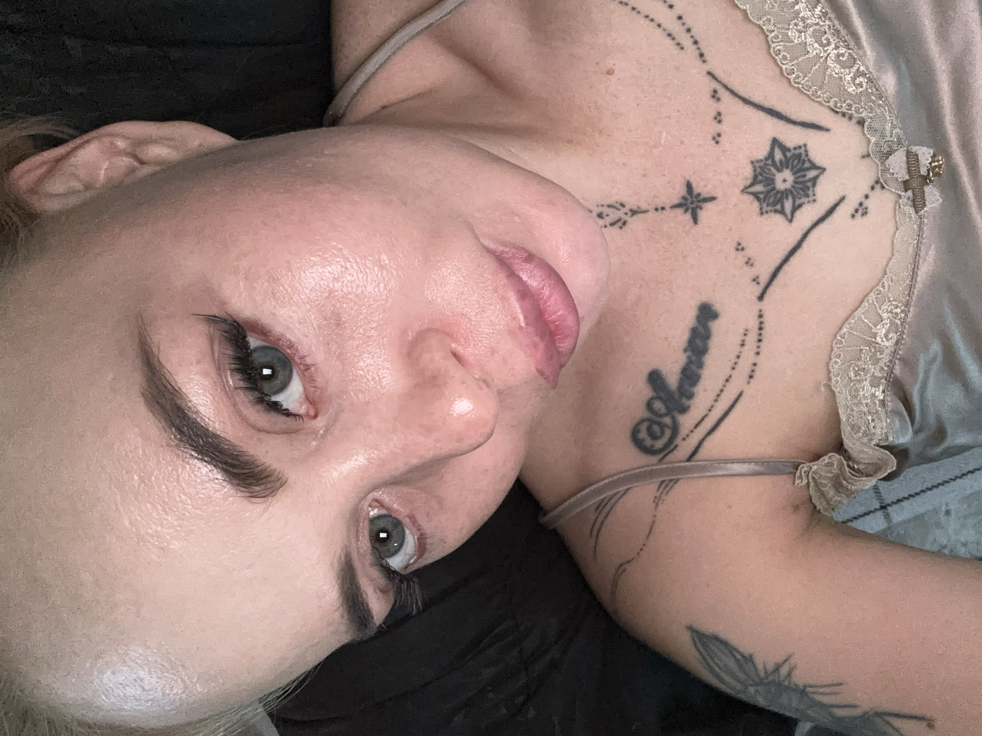 A close-up, high-angle, eye-level shot shows a pale-skinned woman with blonde hair pulled back, looking directly at the camera. She has light blue eyes and is wearing dramatic false eyelashes. Her skin appears slightly shiny and there are visible pores. She has dark, arched eyebrows and her lips are plump and lightly colored. A tattoo is visible on her chest, featuring script, a star, and a floral design, as well as another tattoo on her upper left arm. She is wearing a light-colored, silky camisole with lace trim. The background is dark and out of focus.