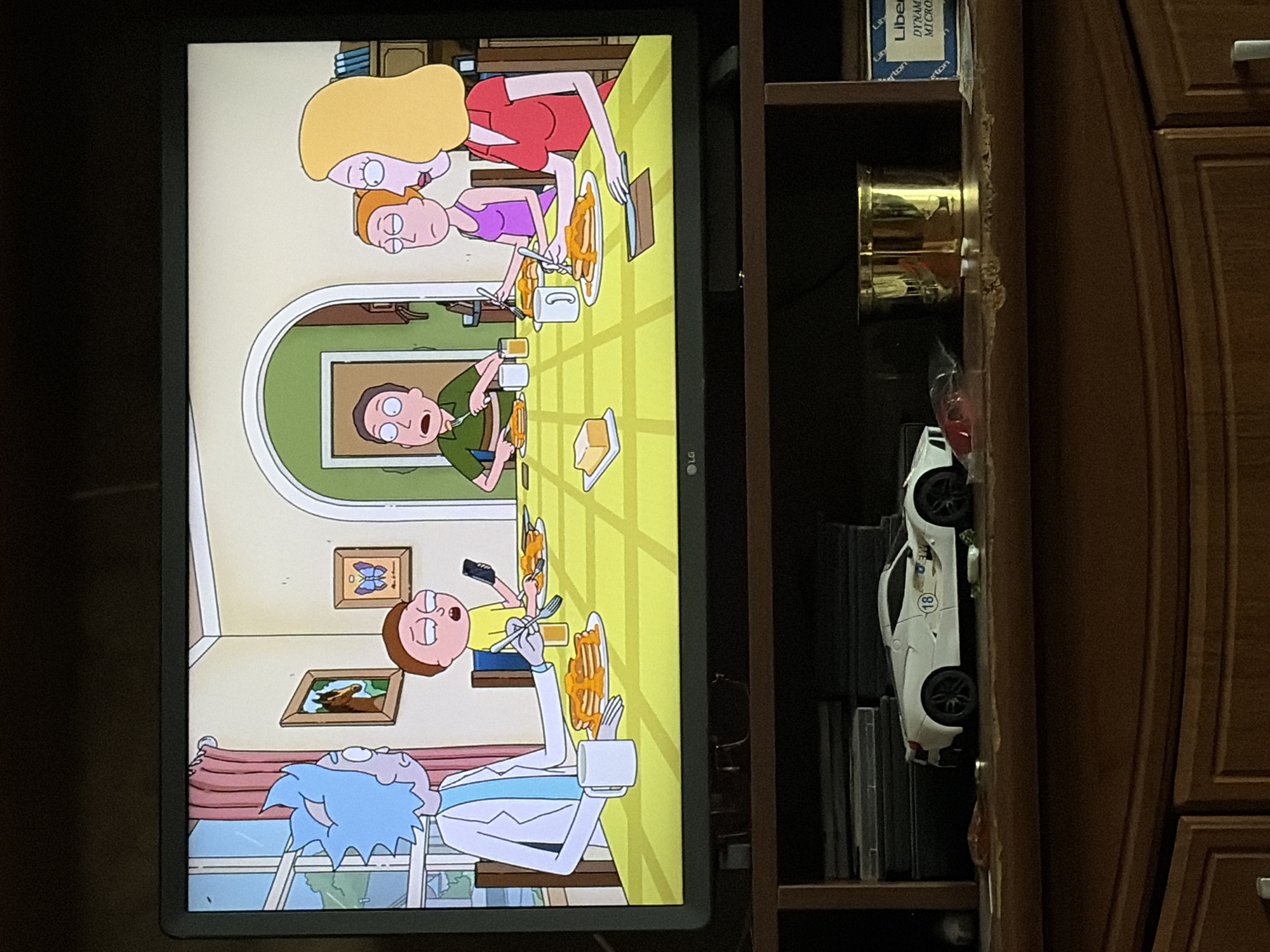 An animated scene depicts a family of four, presumably Rick and Morty characters, at a dining table laden with pancakes and syrup. Rick, with his signature blue spiky hair, wears a lab coat and holds a fork. Morty, in a yellow shirt, looks down at his phone. A woman with blonde hair in a red shirt and a younger girl in a purple tank top are also present, engaged in eating. The room features a window with pink curtains, framed pictures on the wall, and arched doorways. Below the television displaying the animated scene, a shelf holds a white toy car, a gold container, and some boxes.