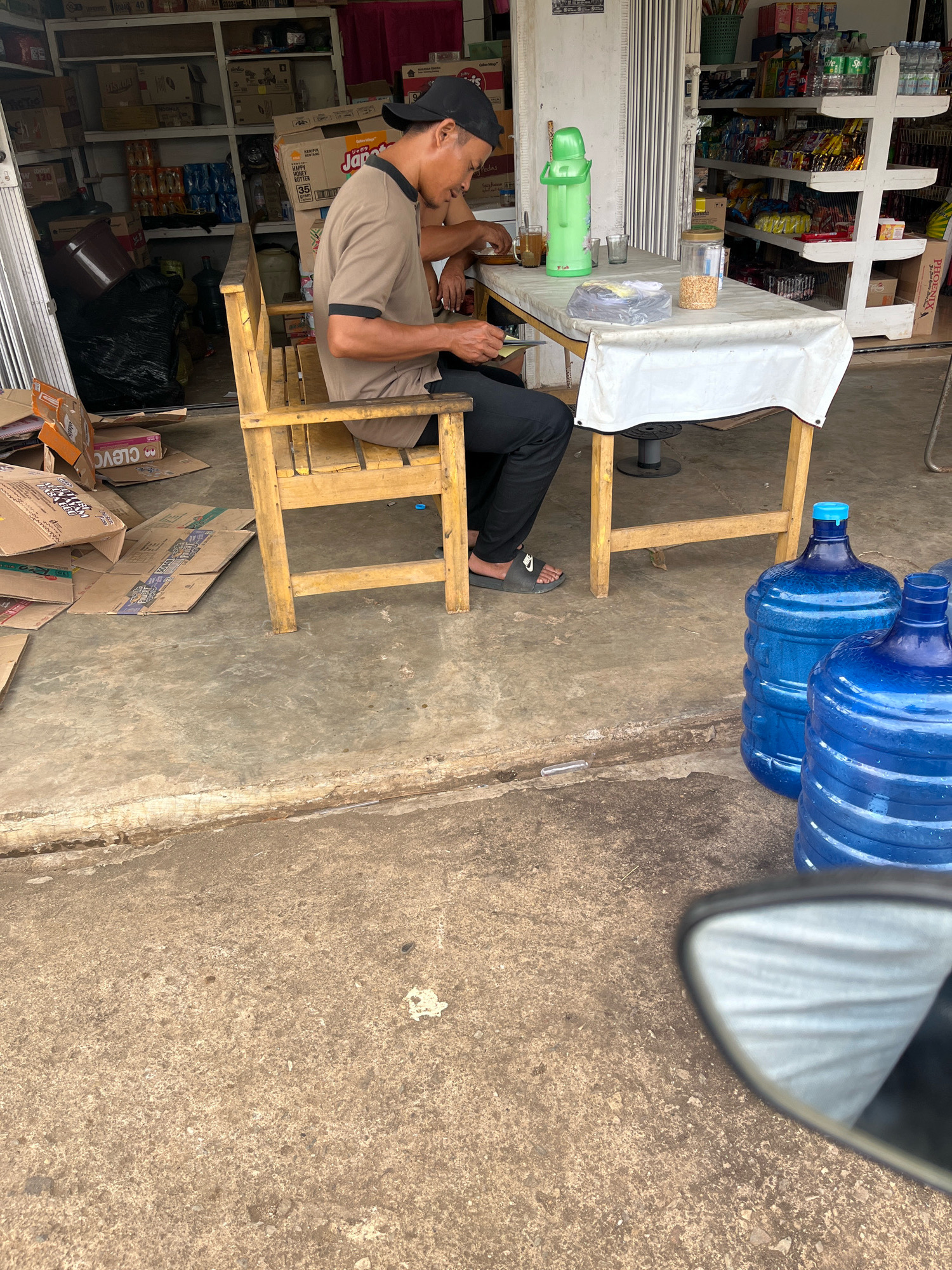 A man wearing a brown shirt, black pants, and a black cap sits on a wooden chair at a table covered with a white cloth in front of a shop. He is looking down and appears to be holding a piece of paper or a card. On the table are a green thermos, two glasses, and a clear container filled with what looks like grain or seeds. Behind the man, shelves stocked with various items are visible, including bottles of drinks and boxes. Two large blue water jugs are on the ground to the right of the table. The scene is outdoors, on a concrete surface, with several cardboard boxes scattered on the ground to the left of the man. The reflection in a motorcycle's side mirror is visible in the lower right corner of the image. The setting appears to be a small store or warung in Muara Dua, Indonesia.