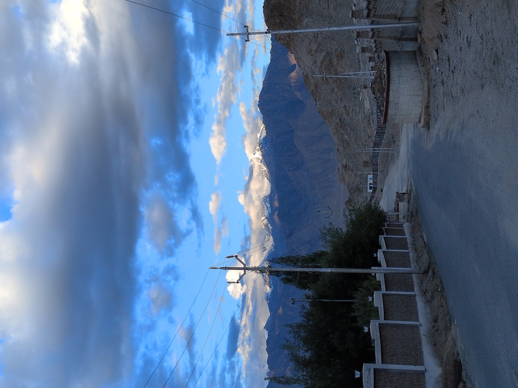 The image depicts a road in the city of Leh, India, with a backdrop of majestic snow-capped mountains. The sky is a dramatic blend of deep blue and dark grey clouds, with patches of white clouds scattered throughout. Power lines stretch across the frame, supported by utility poles. On the left side of the road, a textured wall runs alongside, topped with several modern-looking light fixtures. Beyond the wall, dark green trees create a dense foliage. The road itself appears to be paved and leads into the distance, curving slightly to the right. On the right side of the road, a circular, stone structure stands, its top adorned with rocks. Further down the road, a brick wall with decorative elements lines the other side. The mountainous terrain on the right is barren and rocky, with shadows cast by the uneven landscape. In the distance, the imposing mountain range is a striking feature, its peaks dusted with snow, illuminated by the soft light of what appears to be late afternoon or early morning.