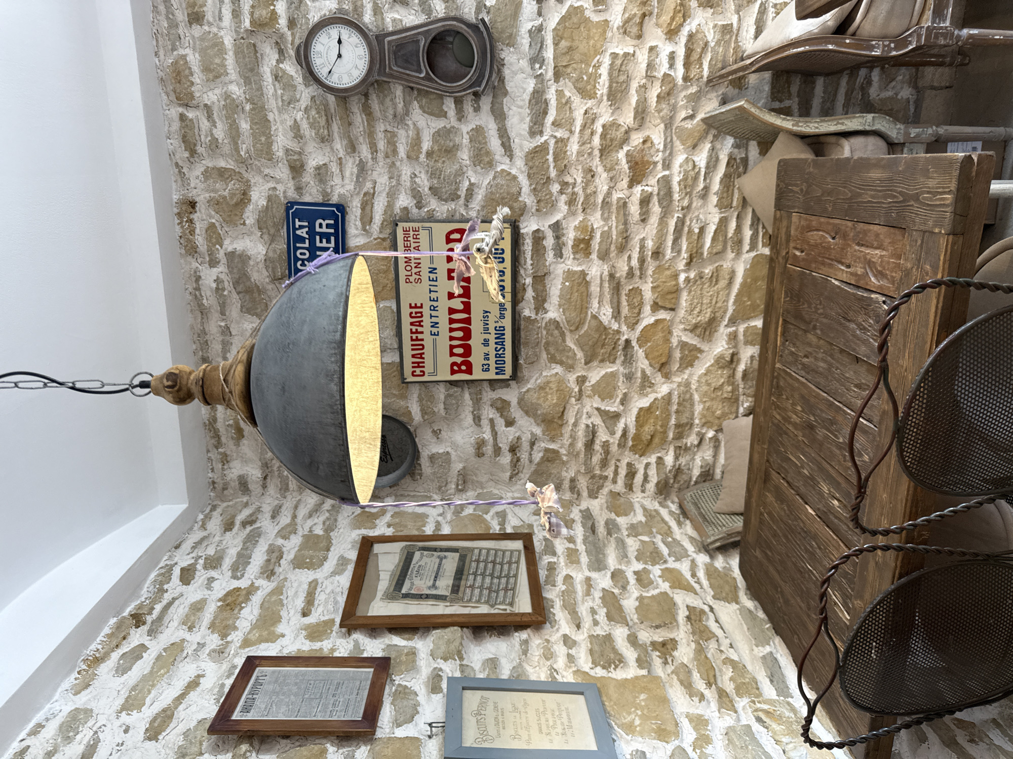The image depicts an indoor scene with a rustic ambiance, possibly a cafe or restaurant, with a prominent stone wall. A wooden table with chairs is centered in the foreground. Above the table, a large industrial-style pendant lamp hangs, casting a warm glow. The stone wall is adorned with several framed items and signs, including what appears to be a vintage clock on the right and a sign with French text that reads "CHAUFFAGE PLOMBERIE ENTRETIEN SANITAIRE BOUILLARD." There are also framed documents and a sign for "BISCUITS PERNOT" on the left. Two chairs with woven seats and metal frames are positioned at the table. The overall environment suggests a cozy and historical setting. There are no people visible in the image. The location in Semogorskiy, Russia is not discernible from the visual cues provided in the image.