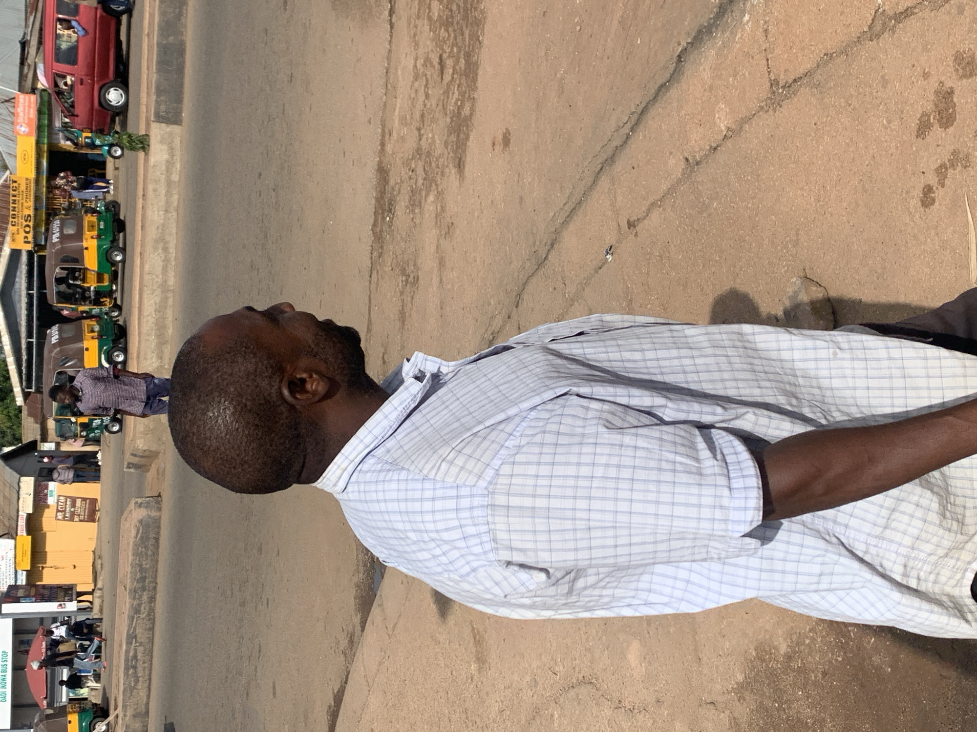 In Gura Top, Nigeria, a man with dark skin and short, dark hair, wearing a light blue and white checkered button-up shirt, stands on a dirt and asphalt road. He is viewed from the right side, looking away from the camera. The road has some cracks and a few dark, wet patches. In the background, there are several vehicles including a red van, two yellow and green tricycles, and a small green scooter. A few other individuals are visible walking and standing near buildings that line the road. One building has signage that includes the words "DADI IKOW A BUS STOP". The scene appears to be a busy street in a commercial area.