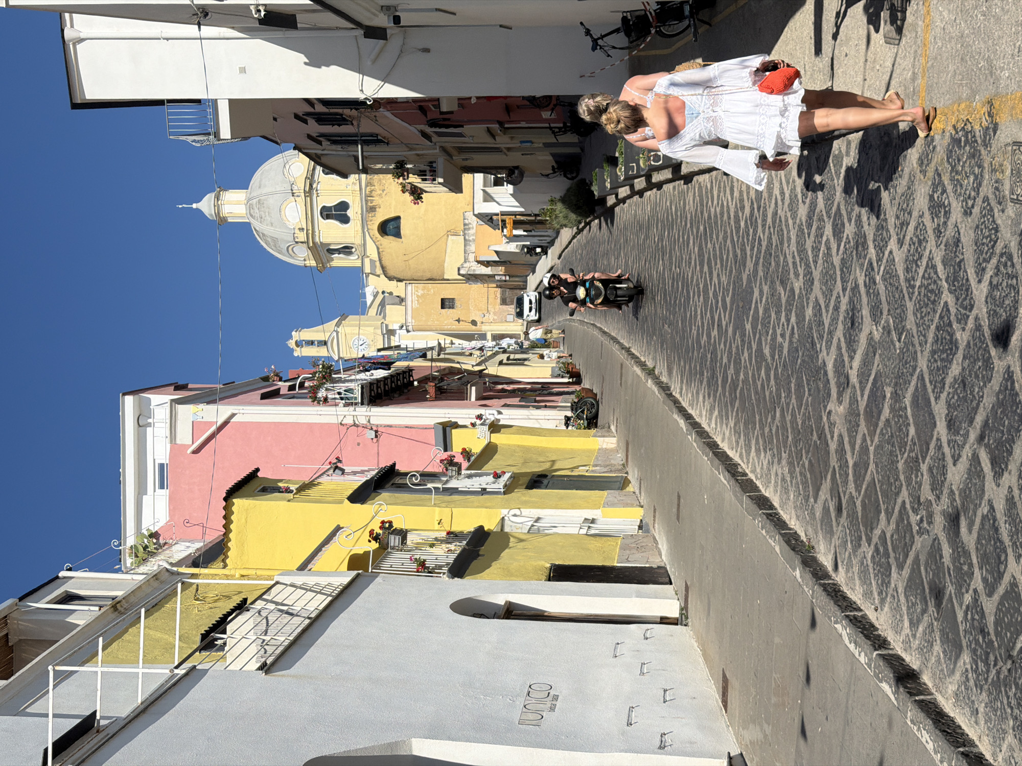 The image depicts a narrow street in Procida, Italy, characterized by colorful buildings. A church with a distinctive dome is visible in the background. Two women, one walking on the sidewalk and another in the street on a scooter, are present. The street is paved with a distinctive pattern. The buildings are painted in shades of yellow, pink, and white. A clear blue sky is visible above.