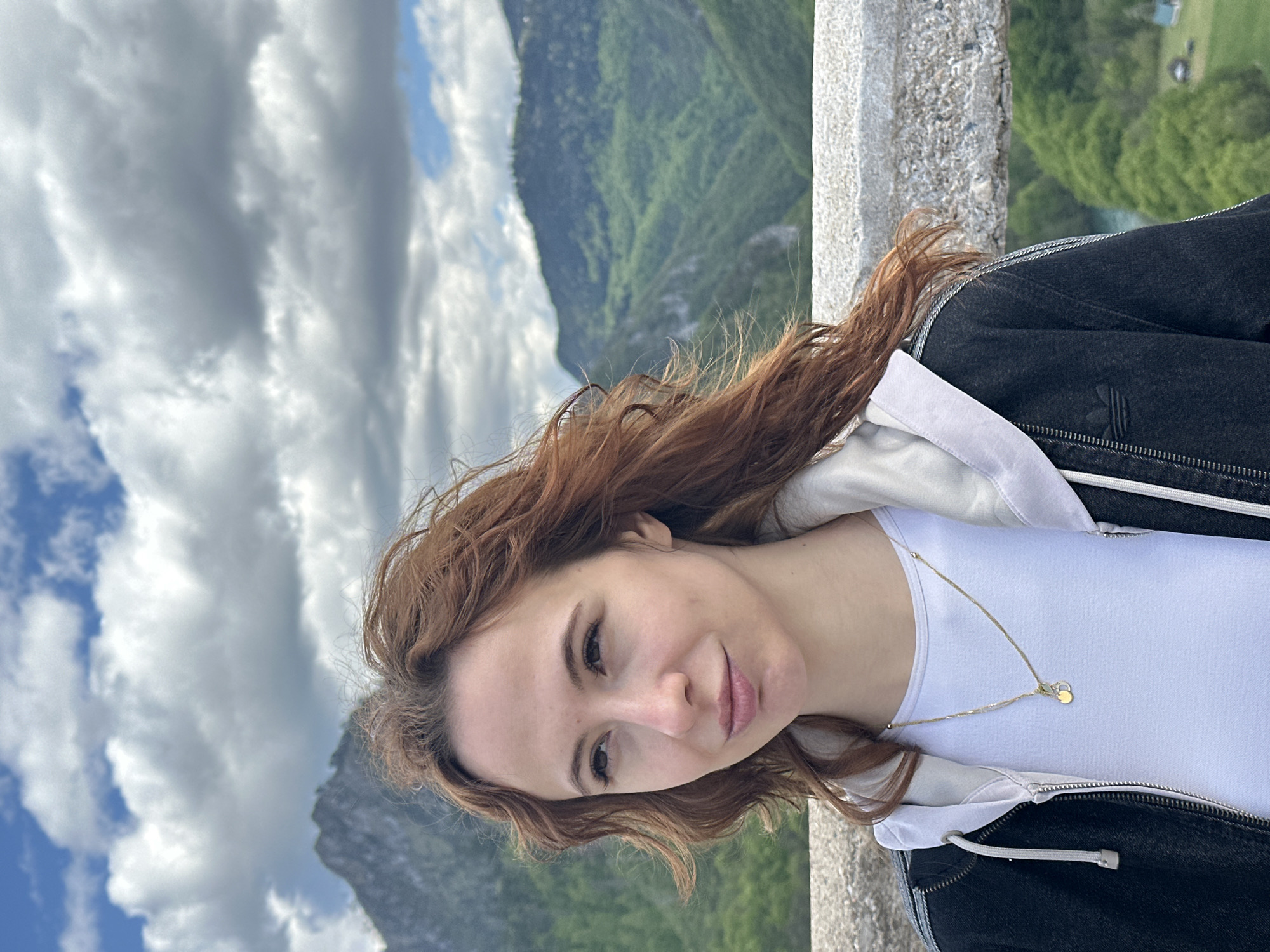 A reddish-haired woman in a white t-shirt and black jacket stands beside a stone railing. Behind her are mountains and a cloudy sky. The location is Đurđevića Tara, Montenegro.