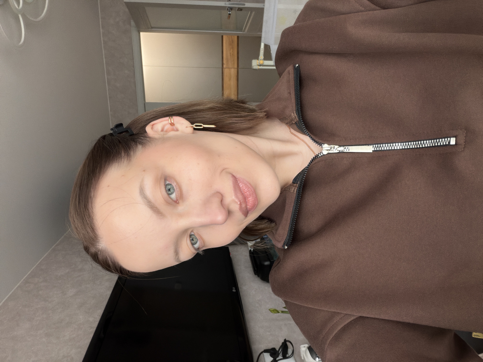 A woman with fair skin and blue eyes, wearing a brown zip-up hoodie and gold earrings, poses indoors. Her hair is brown and pulled back with a black claw clip. A television is visible in the background along with a light-colored wall and a patterned surface. Other items, including glasses and a charger, are also present. The location is Reshetnikovo, Russia.