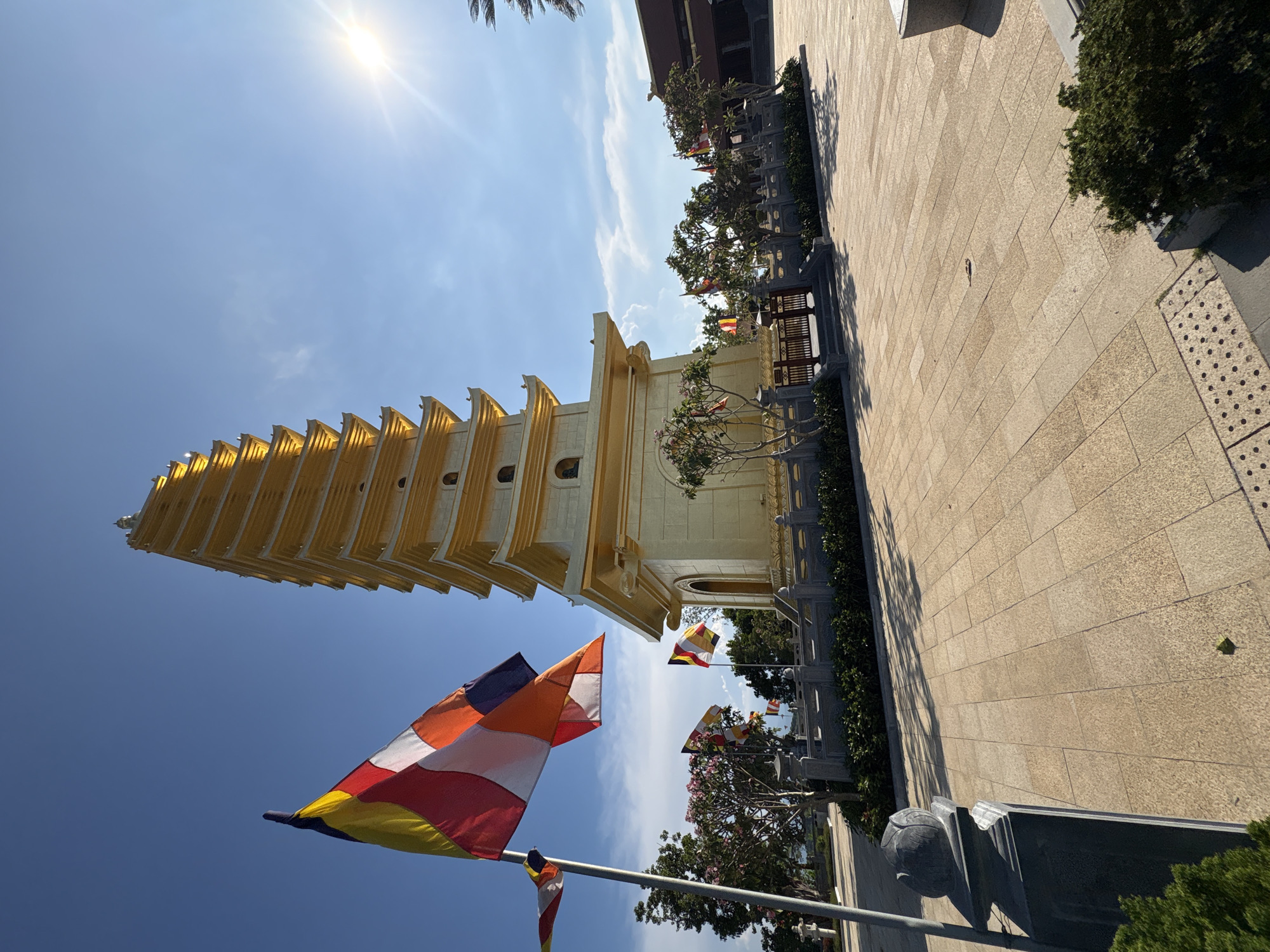 The image shows a tall, golden pagoda located in Diên Khánh, Vietnam. The pagoda has multiple tiers and a distinctive architectural design. In the foreground, several Buddhist flags with horizontal stripes in red, yellow, white, orange and blue are waving in the wind. The sky is bright blue with a few clouds and the sun shining at the upper center. To the side of the pagoda, there is a paved walkway lined with trees and shrubs.