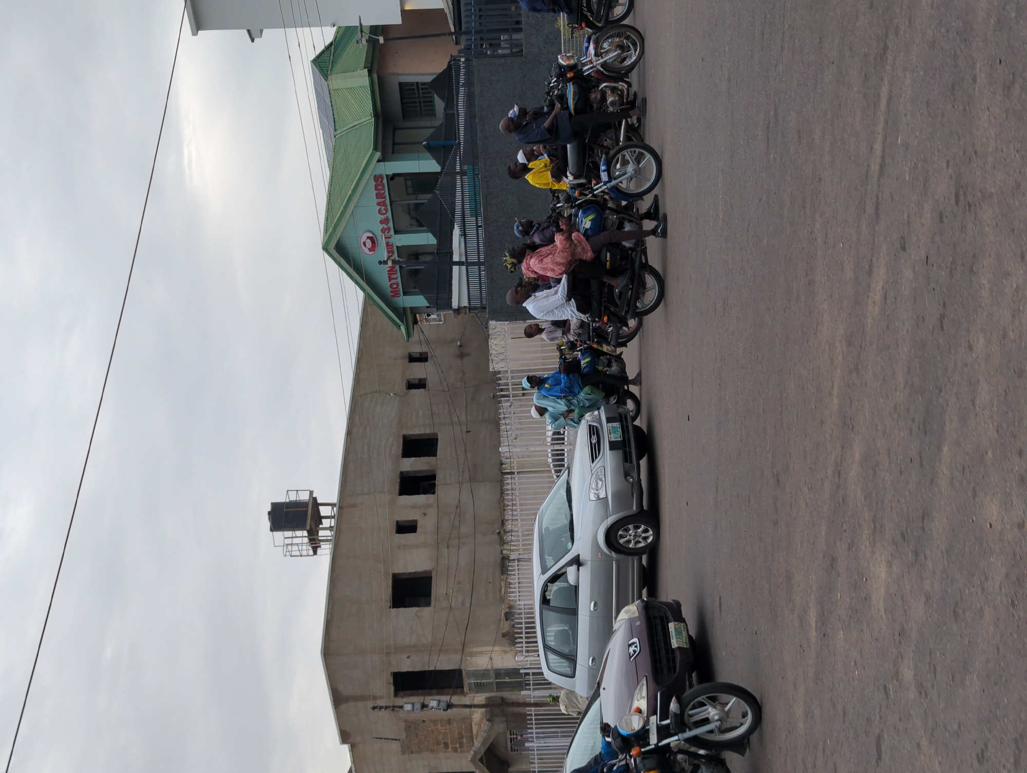 A street scene in Kabala, Nigeria. Multiple motorcycles are parked along the road, some with riders. Several cars are also parked near the motorcycles. Buildings of varying architectural styles are visible in the background. The sky is overcast.