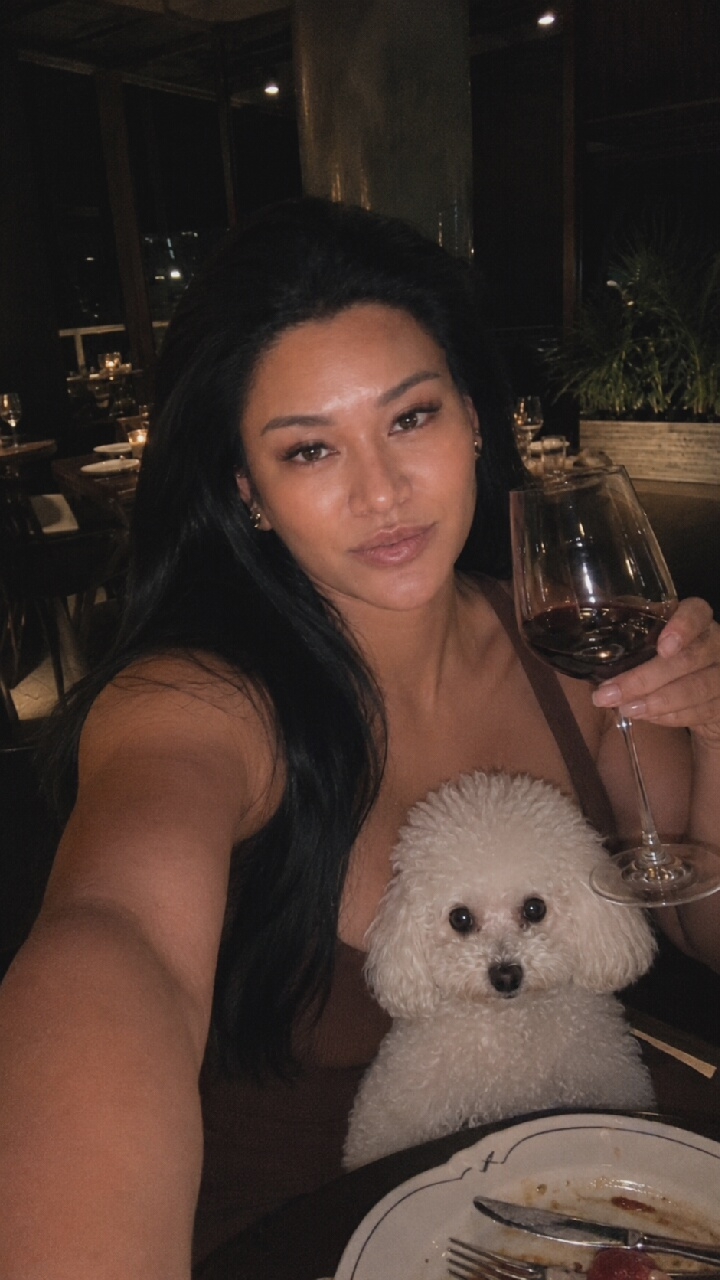 A woman with long, dark hair is seated at a table, holding a glass of red wine. She is looking directly at the camera. A small, white poodle is resting on her lap. The background appears to be a dimly lit restaurant with tables and chairs visible. It is nighttime and the scene suggests an urban setting with city lights visible through a window in the distance. The location is not determinable from the image.