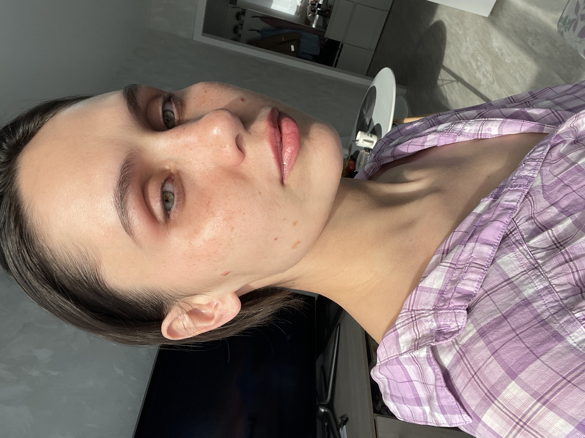 A young woman with fair skin and light brown hair is shown in a close-up, slightly angled portrait. She has green eyes, freckles, and appears to be wearing lip gloss. She is wearing a purple and white plaid shirt. A black object is visible in the background. The location of the image is not discernible.
