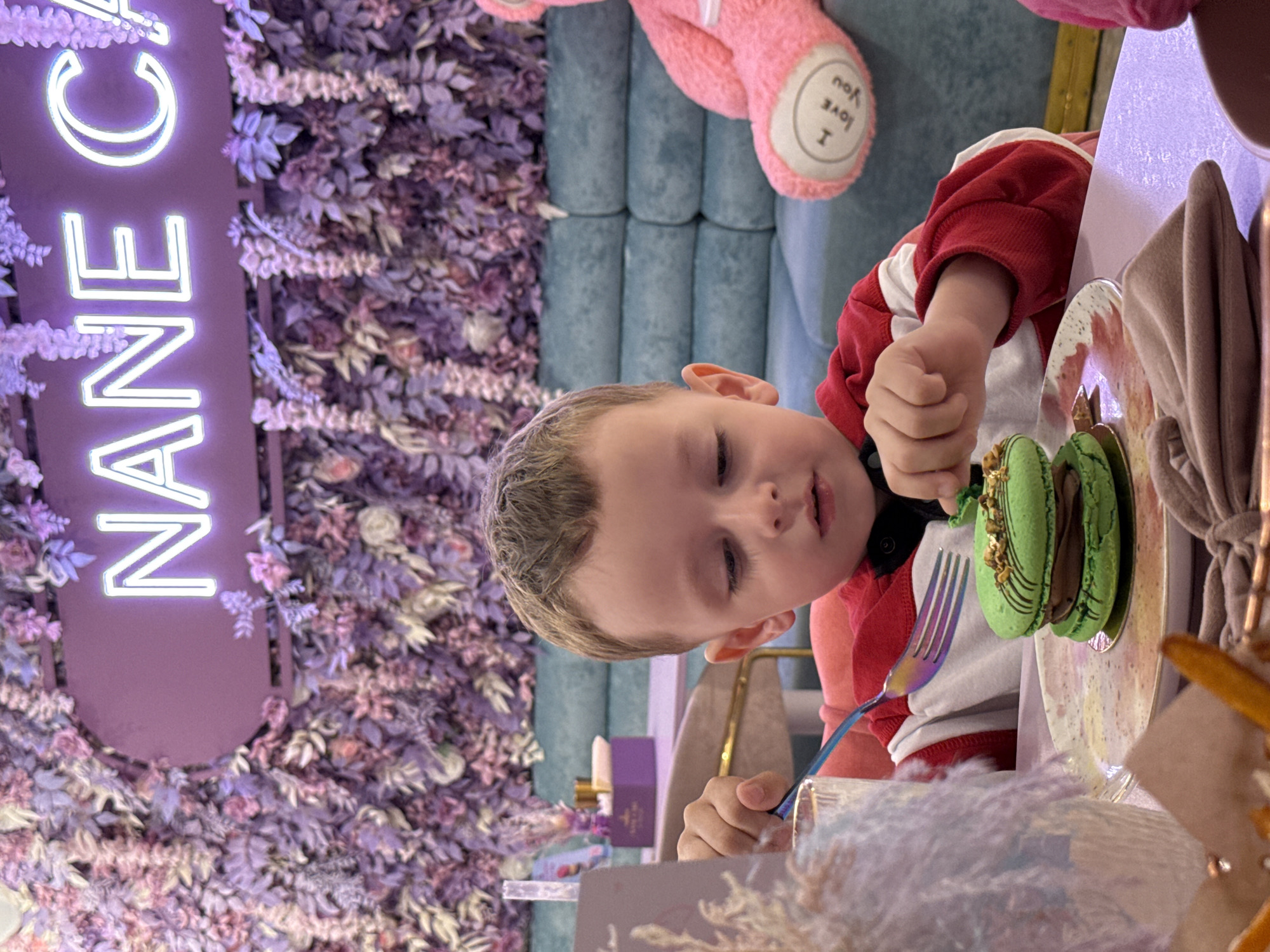 A young child wearing a red and white striped shirt is seated at a table, using a fork to eat a large green macaron. The setting appears to be a cafe or dessert shop. In the background, there is a flower wall with a neon sign that reads "NANEC." A large pink teddy bear with the words "I Love You" on its foot is visible on a blue tufted sofa. The photograph was taken in Vladivostok, Russia.