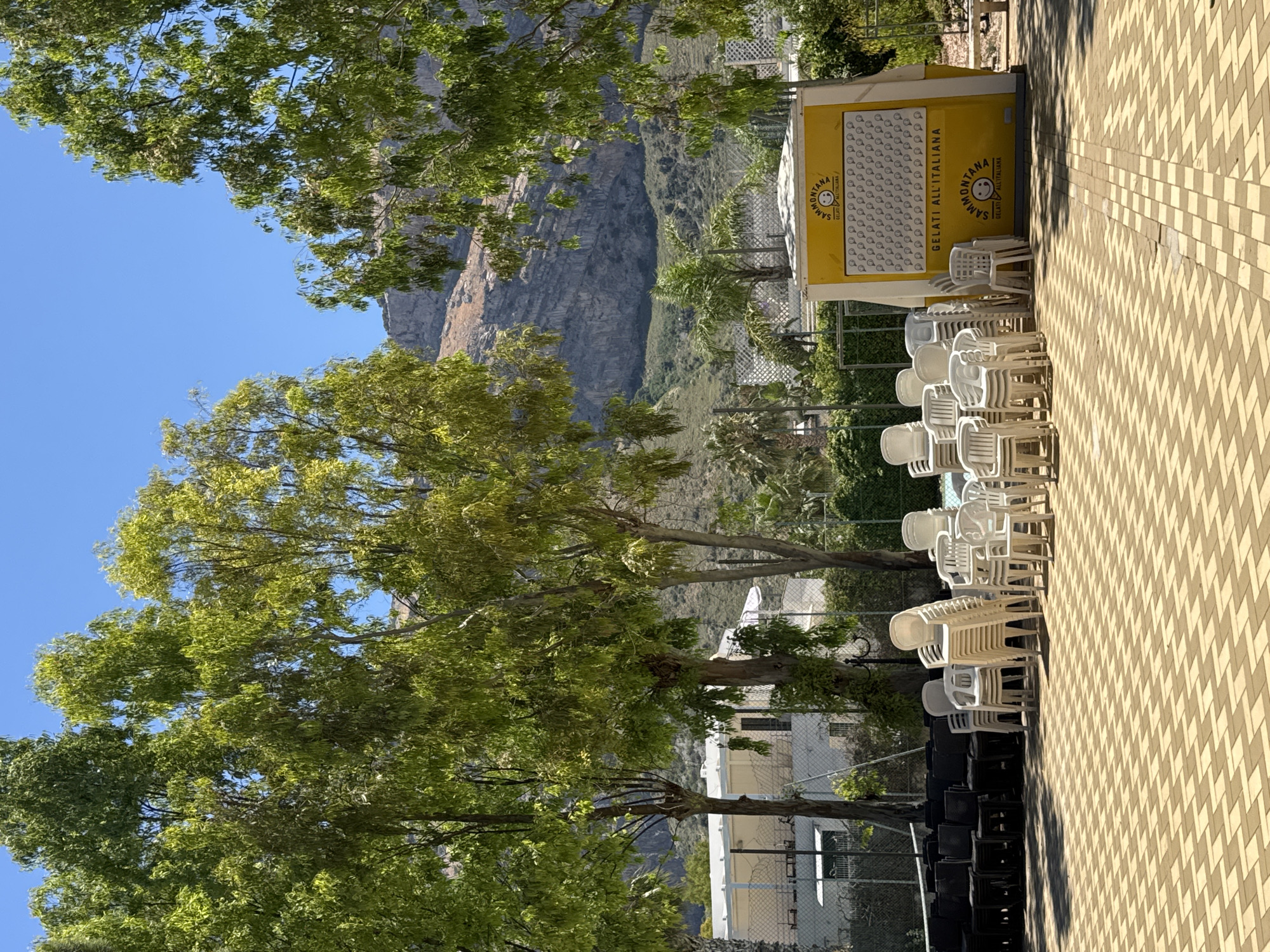 The image depicts an outdoor scene in the city of Isola delle Femmine, Italy, likely in a public square or recreational area. In the foreground, a patterned paved surface is visible, with a large stack of dark chairs on the left and numerous white plastic chairs stacked and arranged to the right. A bright yellow kiosk with the branding "SAMMONTANA GELATI ALL'ITALIANA" is prominently featured on the right side of the image, displaying a grid of ice cream cone graphics. Behind the chairs and kiosk, a fence encloses what appears to be a sports court. Further in the background, lush green trees with dappled sunlight filtering through their leaves dominate the mid-ground. Beyond the trees, a building with a white roof and a fence is visible. The background of the image is characterized by a steep, rocky mountain slope under a clear blue sky. The scene is bathed in bright sunlight, suggesting a warm, daytime setting. There are no people visible in the image.