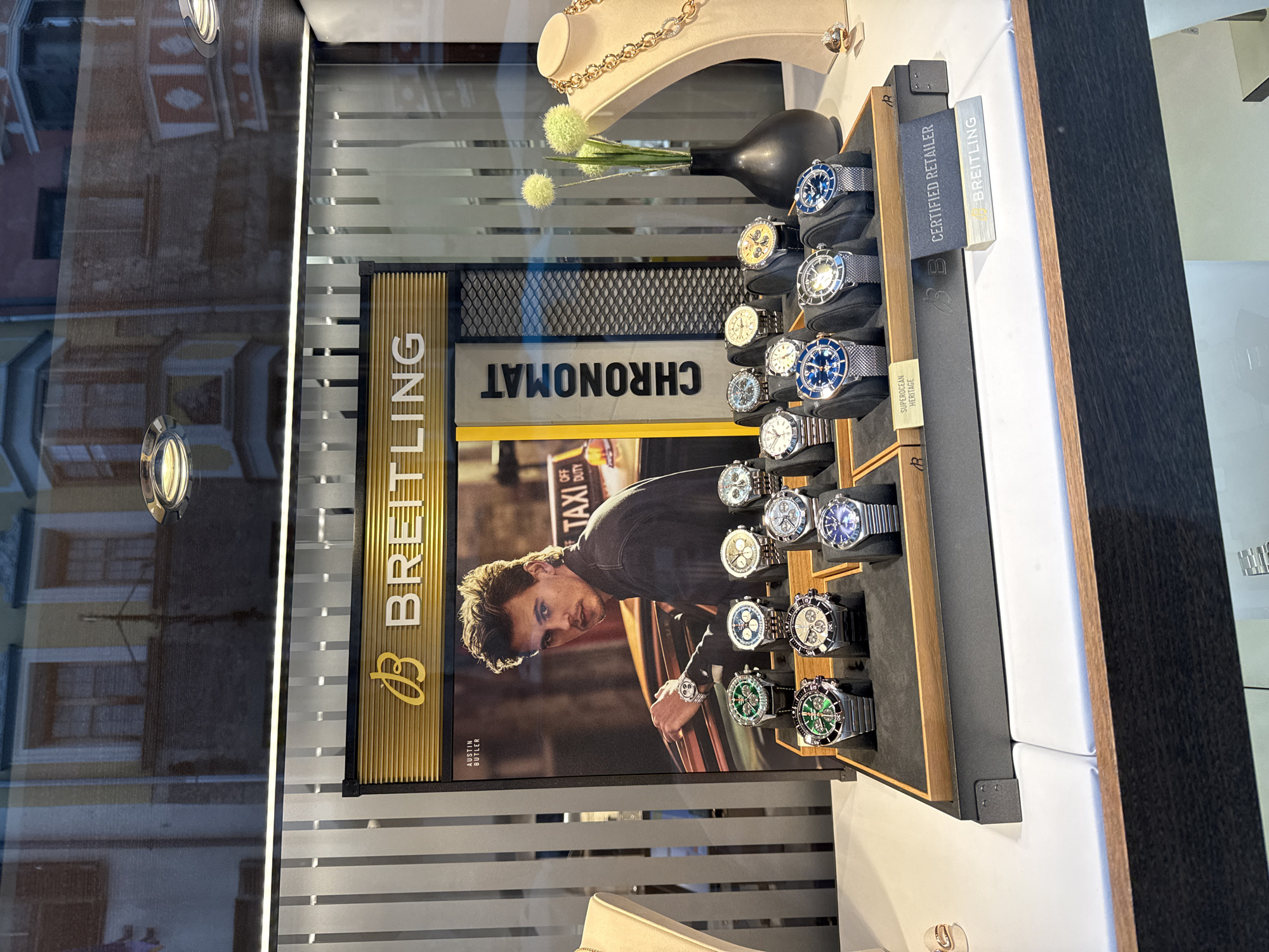 This image shows a Breitling watch display in a shop window, reflecting buildings from Innsbruck, Austria. The display features a large poster of actor Austin Butler with a Breitling watch on his wrist, next to a "TAXI OFF DUTY" sign. Below the poster are several Breitling watches in various styles and colors, arranged on stands. To the right of the watches, a beige jewelry bust displays a gold necklace and a ring. A black vase with three pale green decorative flowers sits behind the watches. A "CERTIFIED RETAILER" sign for Breitling is visible in the foreground. The display is illuminated from above and behind. The reflections in the window show architectural details of buildings, including windows and facades, typical of Innsbruck's historic city center.