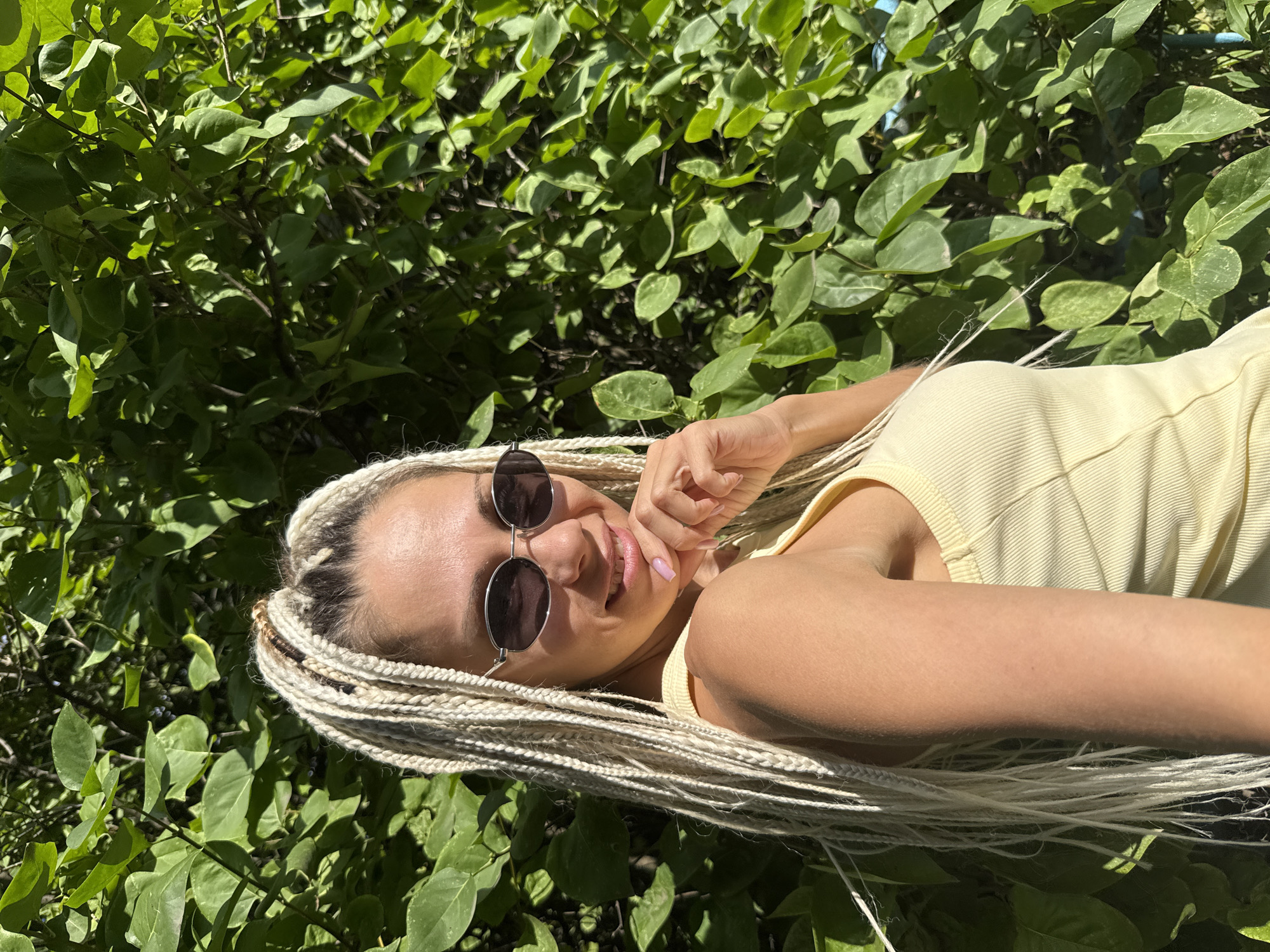 A young woman with blonde braided hair and sunglasses is standing in front of a lush green hedge. She is wearing a light yellow tank top and is smiling, with her hand resting on her chin. The image appears to have been taken outdoors, with natural light illuminating the scene. The background is filled with the dense foliage of the hedge, suggesting a garden or park setting. There is no visible indication that this location is in Rusanovo, Russia.