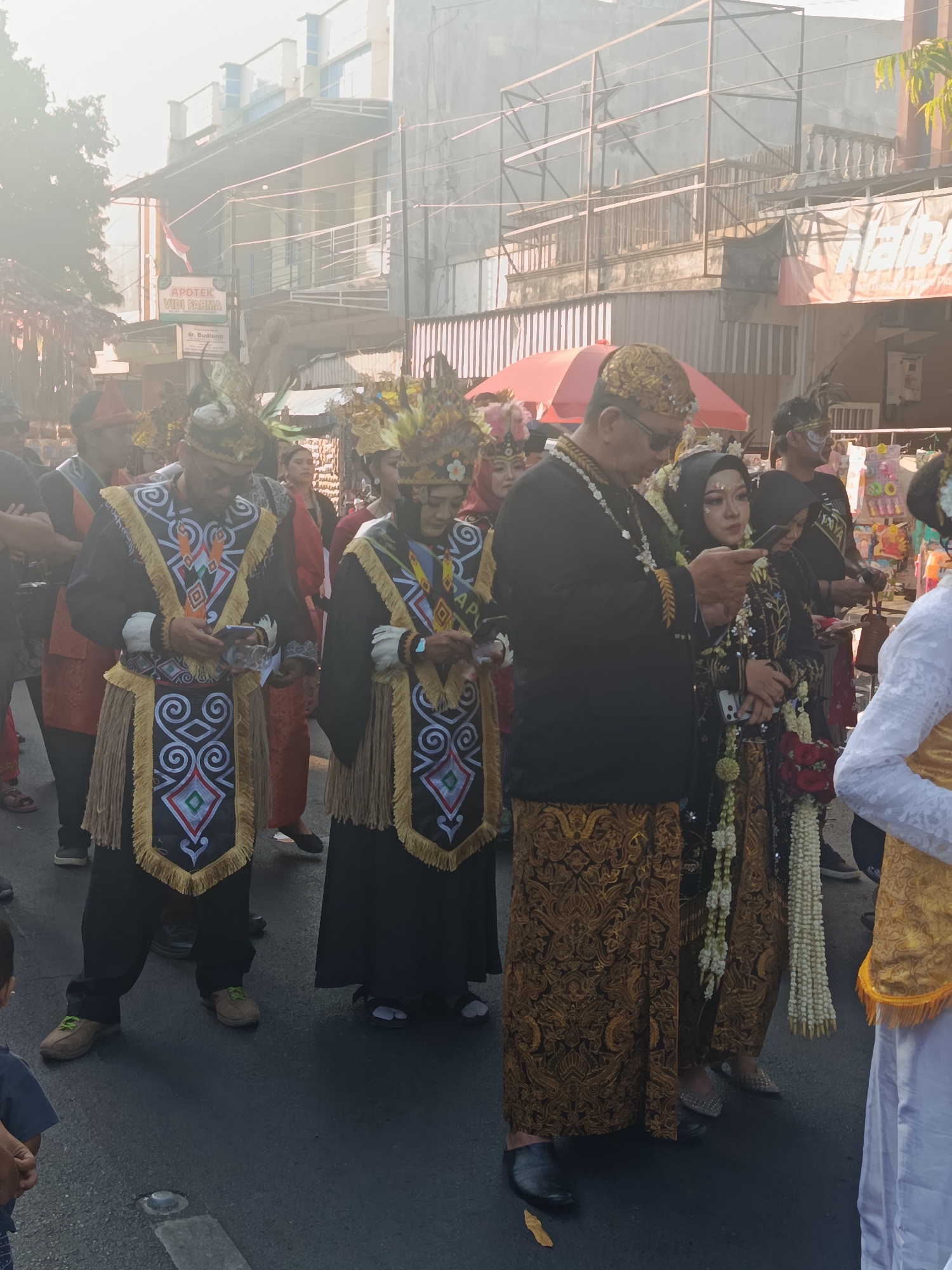 In Ardirejo, Indonesia, a procession is underway on a street. Several individuals are prominently featured, dressed in elaborate traditional attire. Two men in the foreground wear black garments adorned with intricate, colorful geometric patterns on their vests and fringe detailing on their skirts. They also sport ornate headwear. A woman beside them is dressed in a black hijab and a shimmering patterned dress, with a garland of white flowers around her neck, suggesting a ceremonial occasion, possibly a wedding. Other participants in the procession are visible in the background, also wearing traditional costumes and headpieces. The street is lined with buildings, one of which displays signage for "APOTEK VIDI FARMA". The atmosphere appears bright, possibly indicating daytime, with a slight haze or fog present. People are walking, and some are holding objects, potentially for the ceremony.