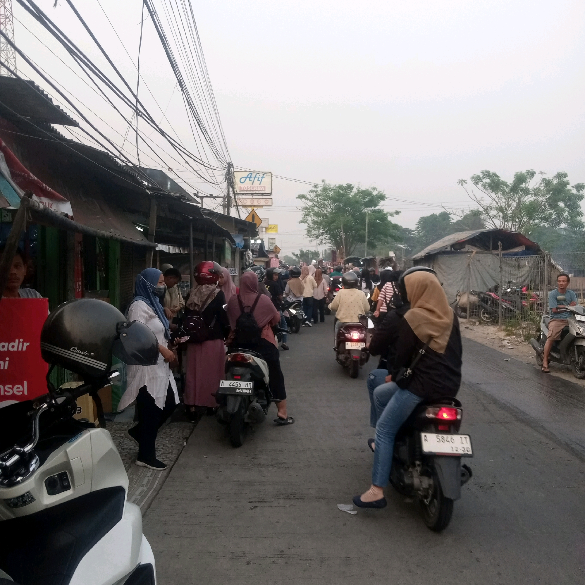 The image depicts a street scene in Nagara, Indonesia, with numerous motorcycles and people. The sky is overcast, suggesting a cloudy day. Along the left side of the road, there are several makeshift stalls or shops with corrugated metal roofs and some awnings. A man is visible behind a red sign on the far left.
A dense crowd of people, many wearing hijabs and casual clothing, are seen on and around motorcycles. Several motorcycles are in motion or parked, indicating traffic or a busy marketplace. The road surface is asphalt.
In the foreground, a white and black scooter is parked. Further down the road, more motorcycles and people are visible, with some individuals riding them. On the right side of the road, a man in a blue t-shirt and shorts is sitting on a parked motorcycle. Behind him, there is a makeshift structure covered with a tarp, possibly a dwelling or storage area, enclosed by a fence. Trees are visible in the background, and the overall impression is one of a bustling, possibly semi-urban or rural commercial area.