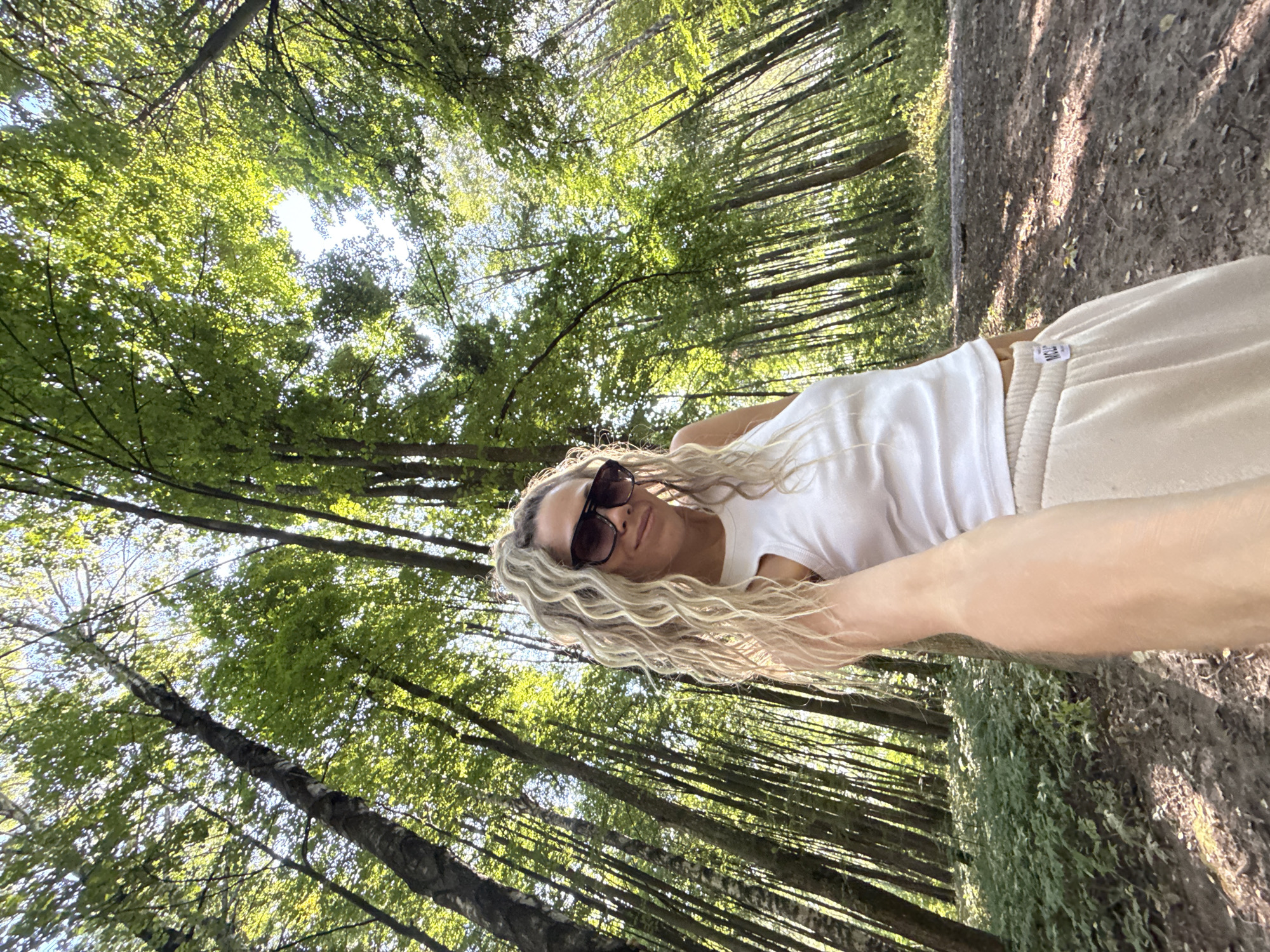 A woman with blonde, curly hair and sunglasses is taking a selfie in a forest. She is wearing a white tank top and beige sweatpants. The trees in the forest are tall and slender, with sunlight filtering through the leaves. The ground is covered in dirt and fallen leaves. The location is specified as Dumino, Russia.
