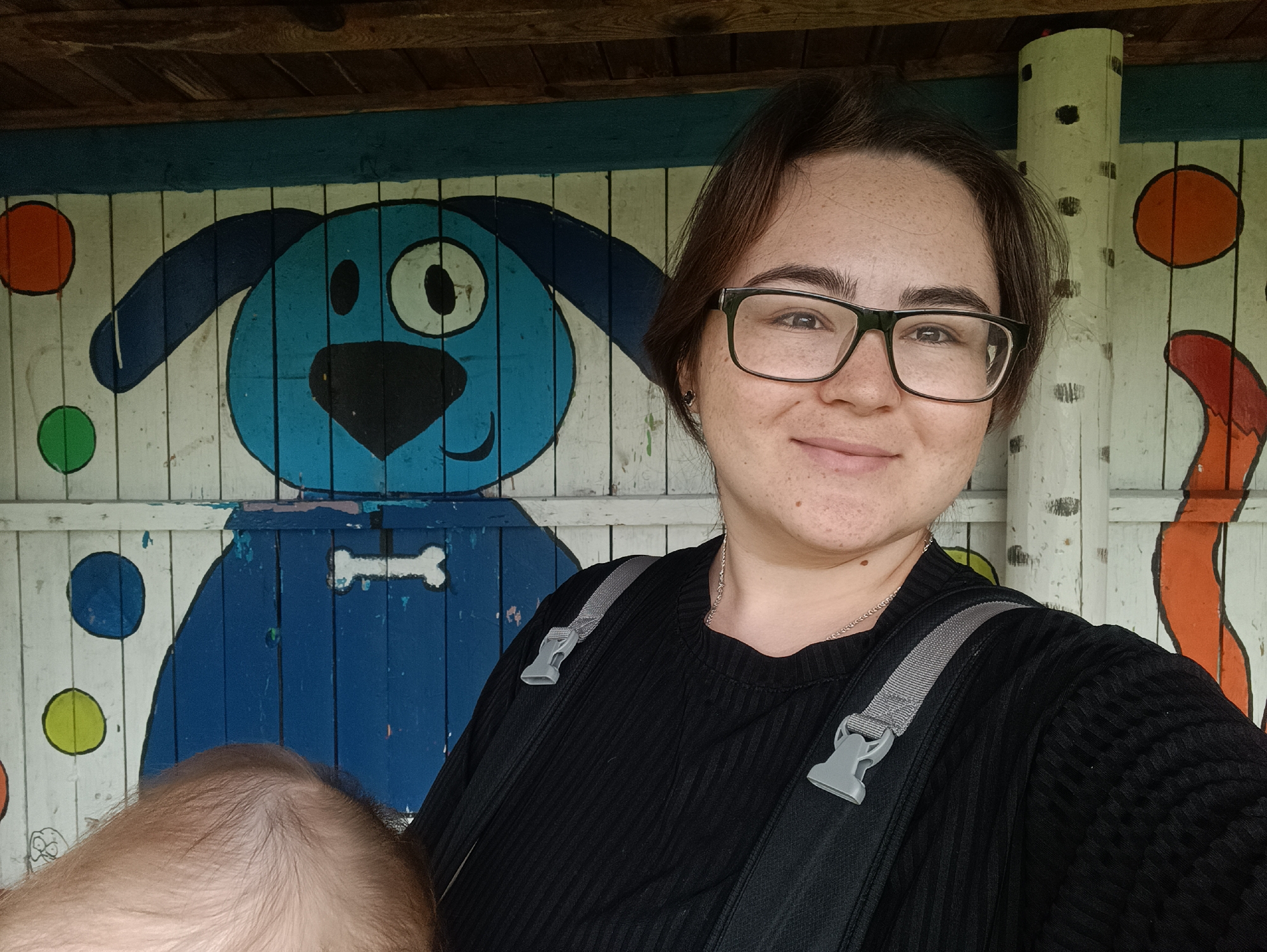 A woman with dark hair and glasses smiles at the camera, holding a baby. The background is a colorful mural painted on wooden planks, featuring a cartoon blue dog with a bone and abstract shapes. Part of the baby's head is visible in the lower left corner. The scene appears to be outdoors, with a wooden roof structure visible at the top. The mural suggests a playful or children's theme, possibly located in a park, playground, or educational facility in the city of Posad, Russia.