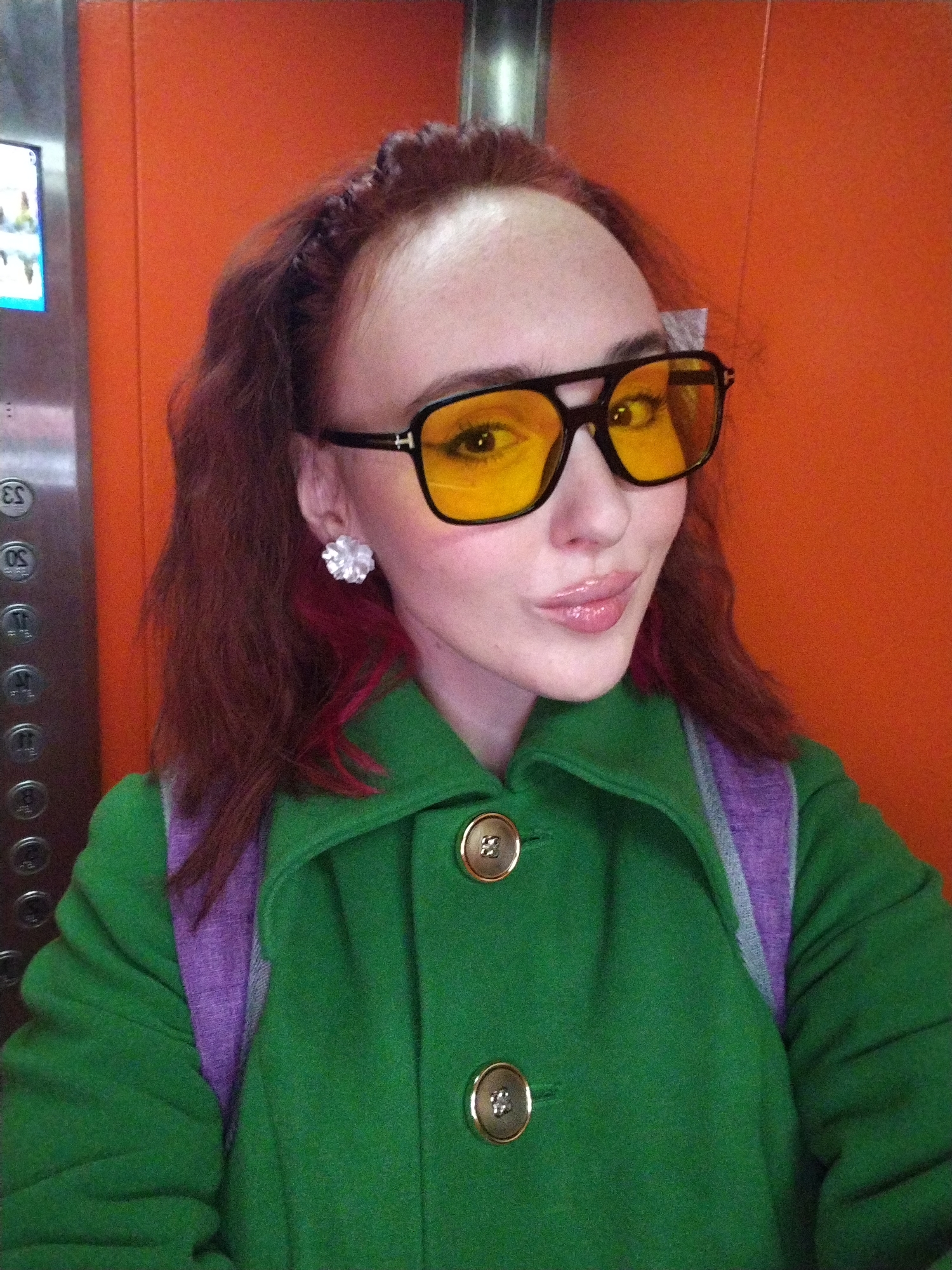 A young woman with reddish-brown hair and yellow-tinted sunglasses poses for a selfie in an elevator. She wears a green coat with gold buttons and a purple backpack strap is visible over her shoulder. She has on a white floral earring on her left ear. The elevator has an orange wall behind her and a panel of numbered buttons on the left side of the frame. The elevator is likely in Izhevsk, Russia.