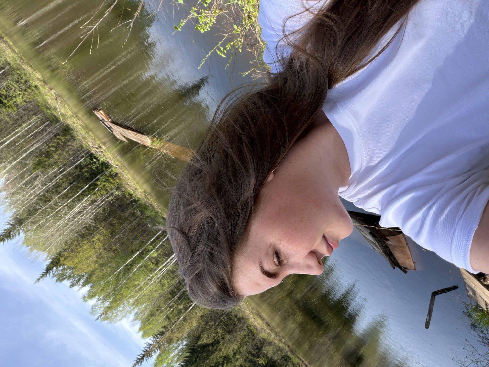 A young woman with brown hair and a white shirt looks down at a body of water. The water reflects the surrounding trees and sky. There is a piece of driftwood floating in the water. Ural'skoe, Russia is not evident in the image. The location cannot be confirmed without additional information.