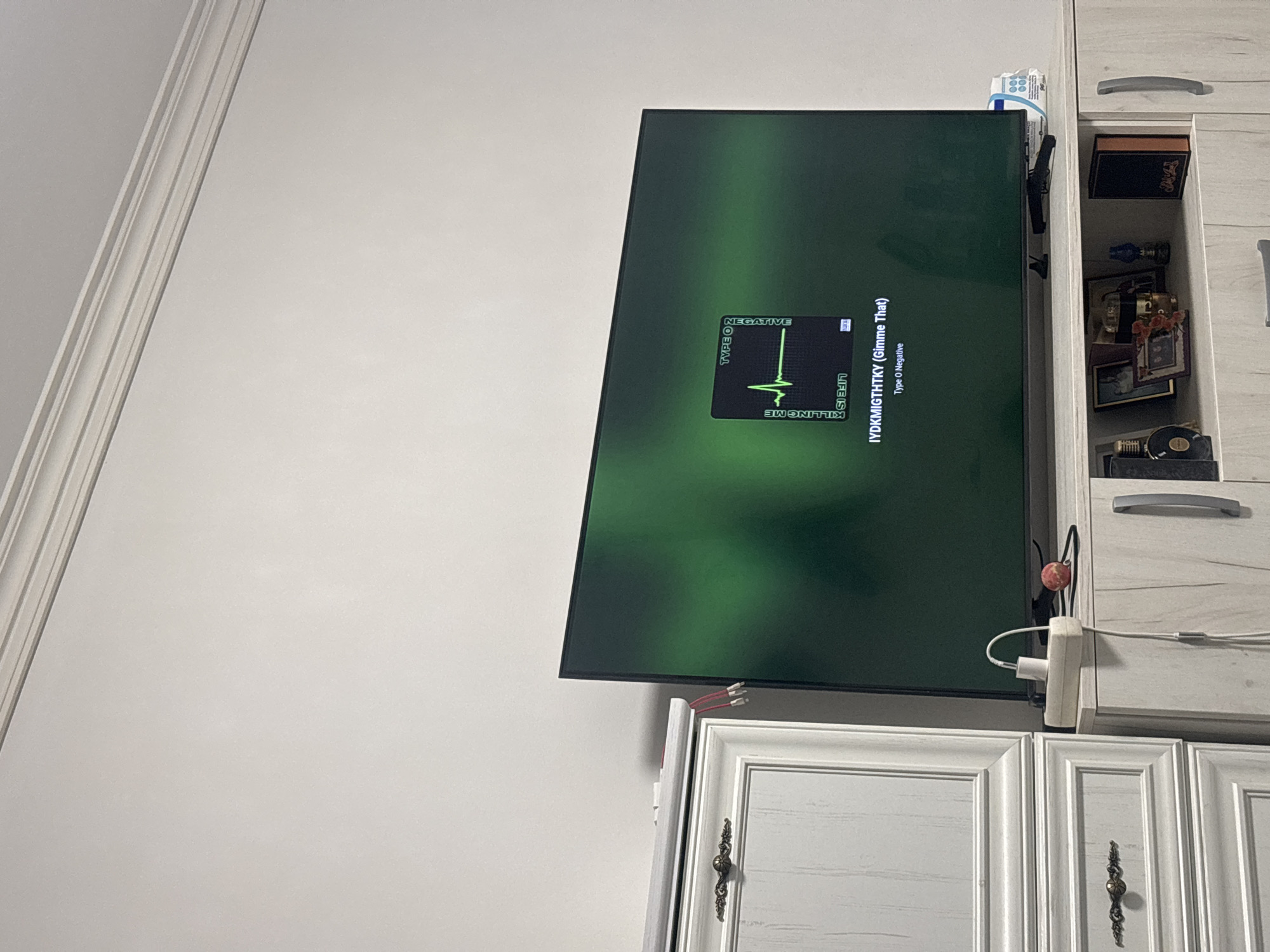 The image shows a large television screen displaying album art for "Type O Negative". The art features a stylized electrocardiogram with the words "KILLING ME", "LIFE IS", and "NEGATIVE" arranged vertically around it. Below the art, the text "IYDKMIGTHTKY (Gimme That)" and "Type O Negative" are visible. The television is placed on top of a light-colored media console. To the left of the television is a tall, white cabinet with decorative molding and a dark, ornate drawer pull. On the media console, to the right of the television, a white power strip with a white charger plugged in is visible, along with a small, round, red object and a white packaged item. The background wall is white and features decorative crown molding at the top. The scene is indoors, likely in a living room or entertainment space. There are no people visible in the image.