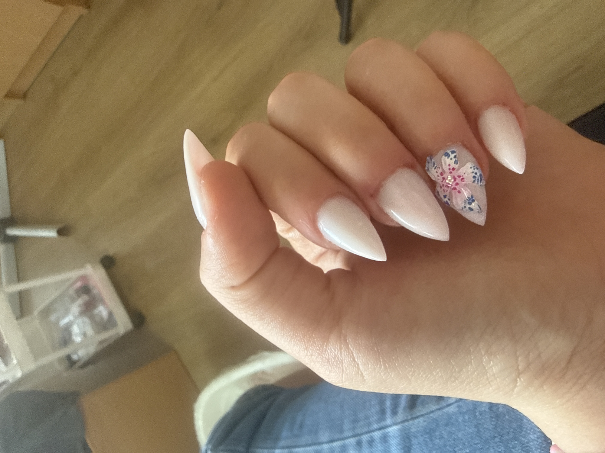 The image presents a close-up view of a person's left hand, angled to display a set of long, pointed (stiletto or almond-shaped) artificial nails. The nails are a uniform milky white or very pale pink color, with a glossy finish. The nail on the ring finger features intricate nail art: a delicate flower design with pink petals, blue accents, and a small, sparkling embellishment at its center.
The hand has a fair skin tone, and the fingers are slightly bent, appearing relaxed. The person is wearing blue denim clothing, visible on the lower right and bottom of the frame, suggesting they are seated. The background is softly blurred, showing a light brown, possibly wooden, floor or table surface. In the lower-left background, a clear plastic container with various indistinct items inside is partially visible. The lighting is bright and appears natural, indicating a daytime setting. There are no specific location cues visible to identify Sofia, Bulgaria, nor is there any visible text. The image focuses entirely on showcasing the detailed nail design.