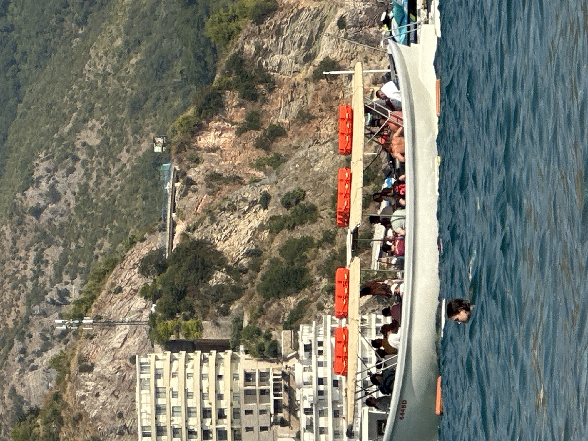 In Boreti, Montenegro, a white tour boat carrying approximately 15 passengers navigates choppy blue waters. The boat features a canvas canopy for shade, with several bright orange life vests stored on top. Passengers are seated and standing, some engaged in conversation. In the foreground, a single swimmer with dark hair is visible in the water. Behind the boat, a large, multi-story white building with numerous windows is nestled against a steep, rocky hillside covered in green vegetation. A cellular tower stands on the upper part of the hill, with a small structure also visible on the cliff face. The scene captures a day of leisure and sightseeing along the Montenegrin coast.