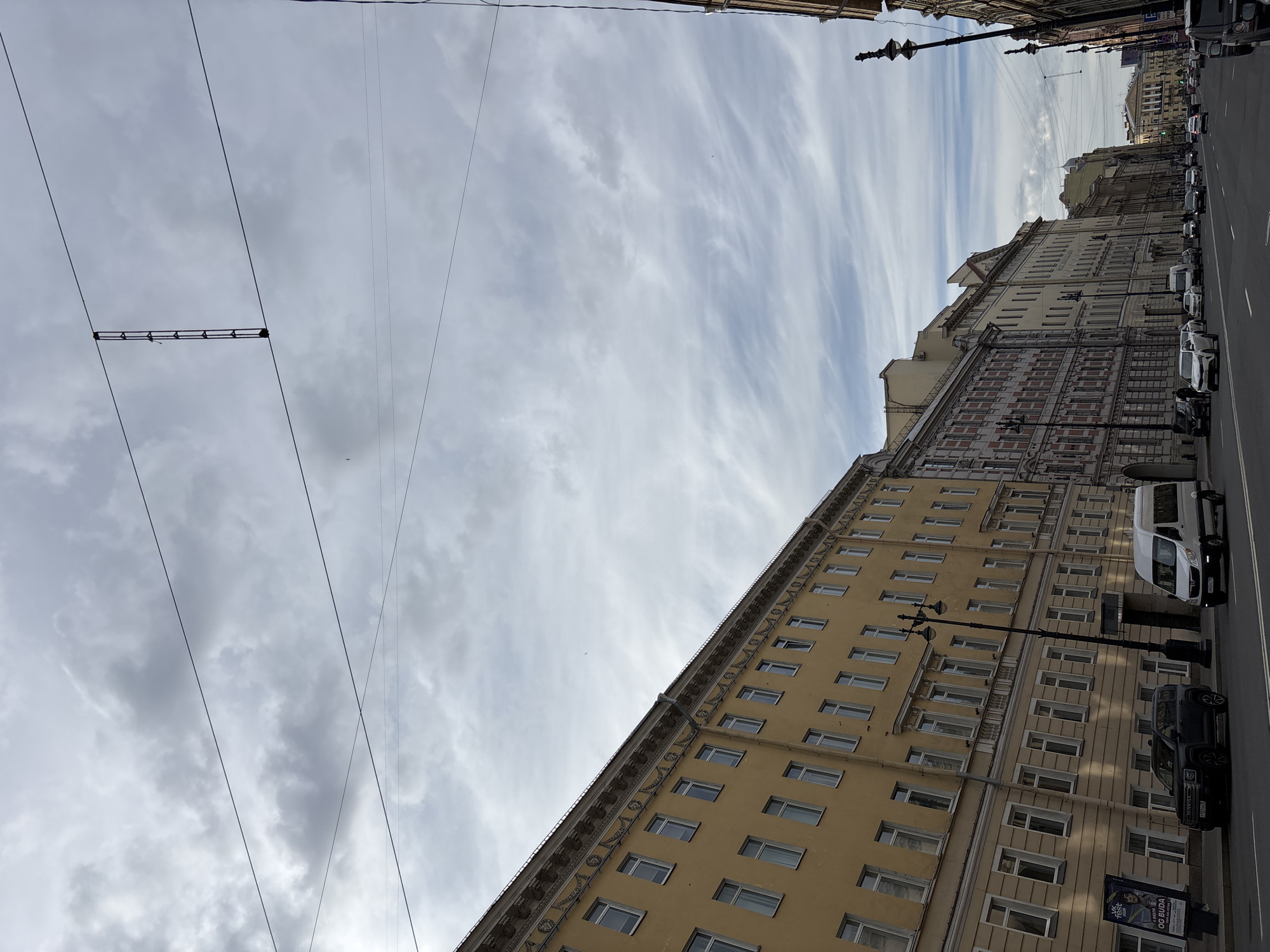 This media file captures a street scene in Saint Petersburg, Russia, under a cloudy daytime sky. The image is composed at an angle, presenting a long stretch of multi-story buildings lining a paved road where cars are parked.
The most prominent building on the right features a light yellow or beige facade with numerous rectangular windows uniformly spaced across its many floors. It exhibits classical architectural elements, including decorative cornices and moldings beneath the roofline and between floors. Further down the street, additional buildings with similar urban European architecture are visible, some with reddish-brown decorative patterns and varying heights.
Along the street, a paved road with white lane markings stretches into the distance. Several vehicles, including a white van and multiple darker cars, are parked at the curb. Ornate black streetlights or lampposts stand at intervals along the sidewalk. A small billboard near the bottom right displays partial text "VK..." and "OG BUDA."
The sky occupies a significant portion of the image, filled with a mix of overcast clouds, ranging from dense, darker gray formations to lighter, wispy streaks, suggesting a partly cloudy to overcast day. Numerous black overhead wires crisscross the sky, likely part of a public transport system such as trolleybuses or trams. One section of these wires includes a black rectangular apparatus with multiple connectors. A tiny dark speck, possibly a distant bird or small aircraft, is faintly visible against the lighter clouds. The overall scene depicts a typical urban environment during the day.