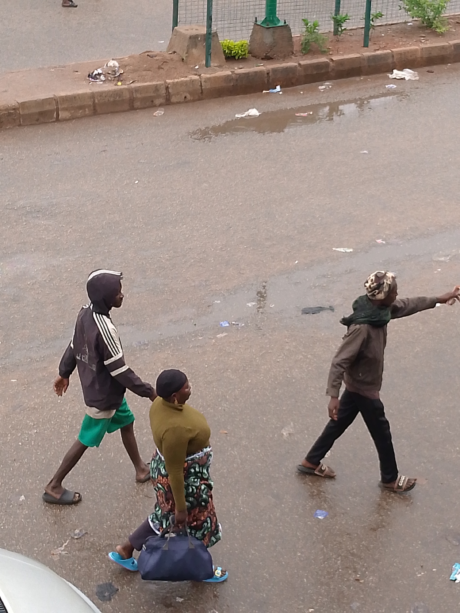 This aerial shot captures a street scene in Bukuru, Nigeria, following a recent rainfall, indicated by the wet asphalt road and scattered puddles. Three individuals are walking on the street.
On the left, a person, possibly a young male, walks towards the left side of the frame. He wears a dark hooded jacket with white stripes characteristic of Adidas on the sleeves and a faintly visible logo on the back, green shorts, and dark sandals.
Next to him, slightly behind, is a woman. She wears an olive-green long-sleeved top and a colorful patterned wrapper or skirt. She is carrying a dark blue duffel bag in her left hand and wears light blue sandals.
Further to the right, another person, likely a male, walks towards the right side of the frame. He is dressed in a brown jacket, dark trousers, and sandals, one of which clearly displays a "GUCCI" pattern. He has a patterned head covering and a green scarf around his neck. His right arm is outstretched, seemingly pointing or hailing something or someone off-camera.
The scene also shows a raised curb with some dirt and sparse vegetation along the side of the road, behind which a green metal fence is visible. Litter, including plastic wrappers, is scattered on the road surface and next to the curb. Part of a light-colored vehicle is visible in the bottom left corner. The lighting appears overcast, suggesting either a cloudy day or early morning/late afternoon.