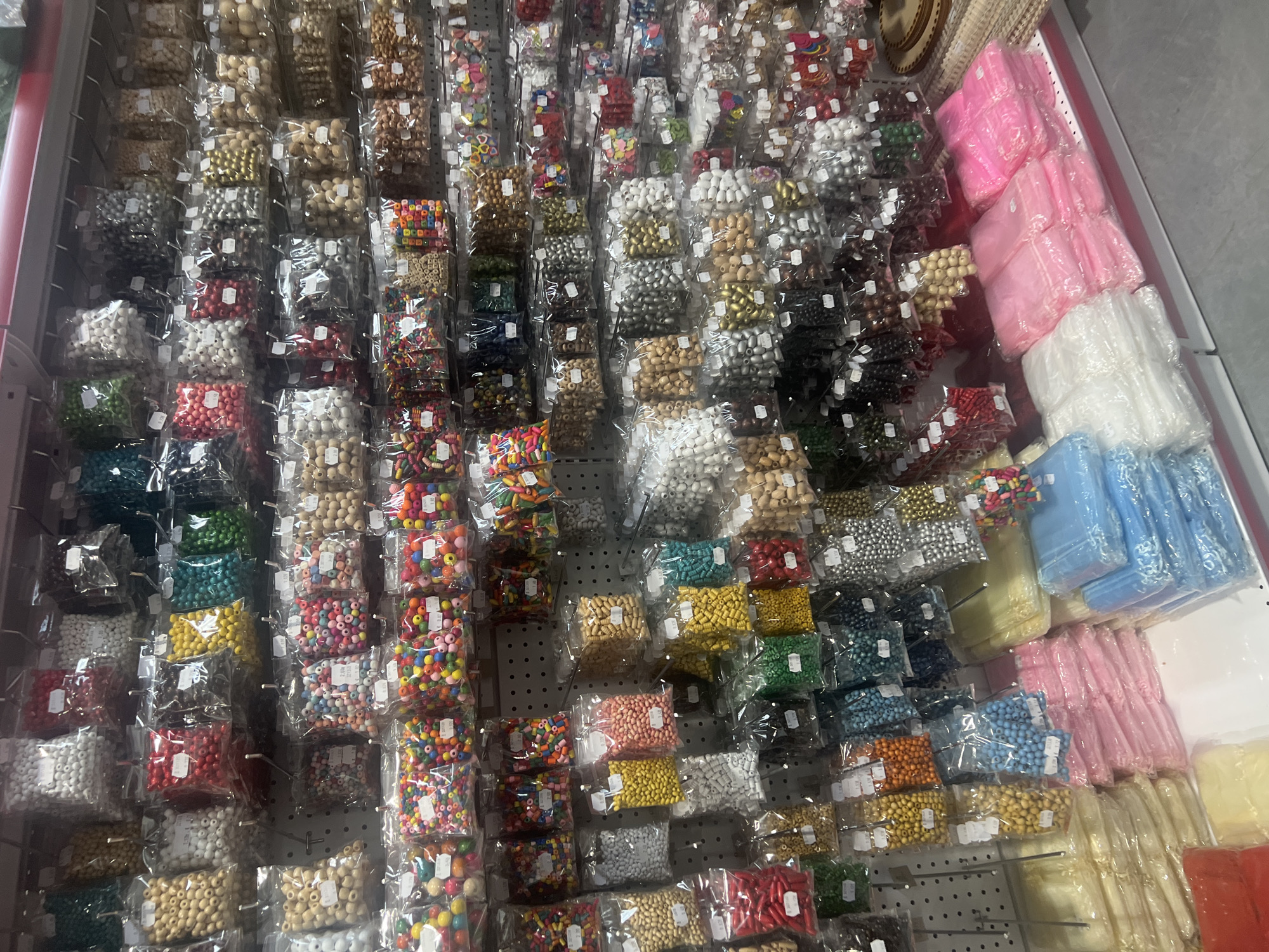 The image shows a display of packaged beads and craft materials, likely inside a shop. The location may be in Burgas, Bulgaria, but this is not verifiable from the image itself.
Rows and columns of small, clear plastic bags are visible, each containing an assortment of beads of varying colors, sizes, and materials (possibly wood, plastic, and metal). Colors include white, black, brown, silver, gold, red, green, yellow, blue, pink, and multicolored.
On the right side of the image are stacks of non-bead items, possibly tulle or similar fabric, packaged in shades of pink, white, blue, yellow, and orange.
The display is attached to a perforated metal wall with hooks, indicating it is a retail setting. The lighting appears to be artificial, casting some shadows within the display. The image is taken from a slightly elevated angle, providing a comprehensive view of the product arrangement.