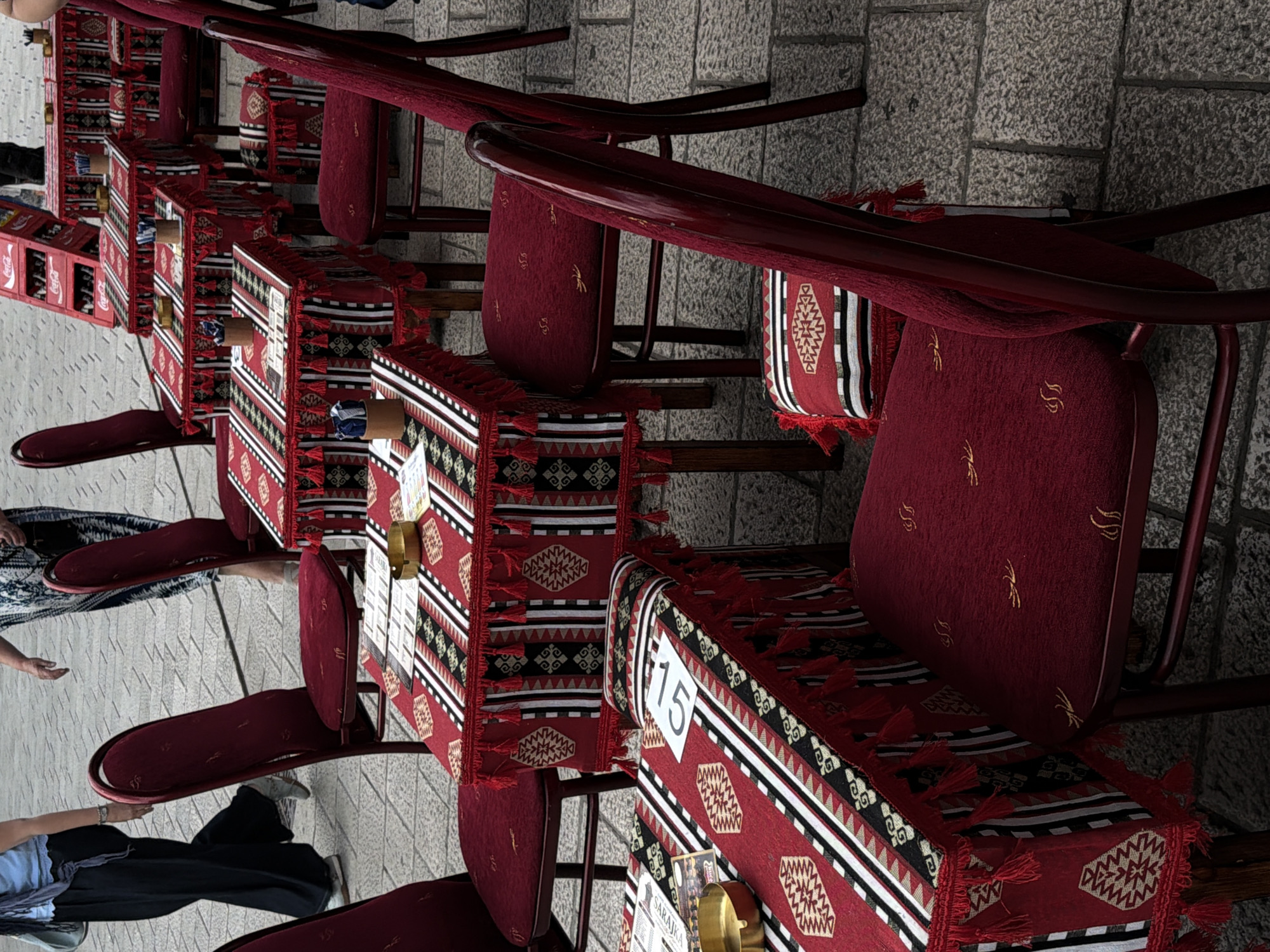 The image shows an outdoor seating area of a cafe or restaurant with tables and chairs set up on a cobblestone street. The tables are covered with red tablecloths featuring a black and white geometric pattern and red tassels. Several chairs are also visible, upholstered in a matching red fabric. Some tables have menus and ashtrays on them. In the background, a red Coca-Cola crate is visible. Two women are partially visible walking in the background, one wearing black pants and a blue top, and the other a patterned dress. The setting appears to be a pedestrian area in Sarajevo, Bosnia and Herzegovina, given the cobblestone pavement and outdoor cafe setup.