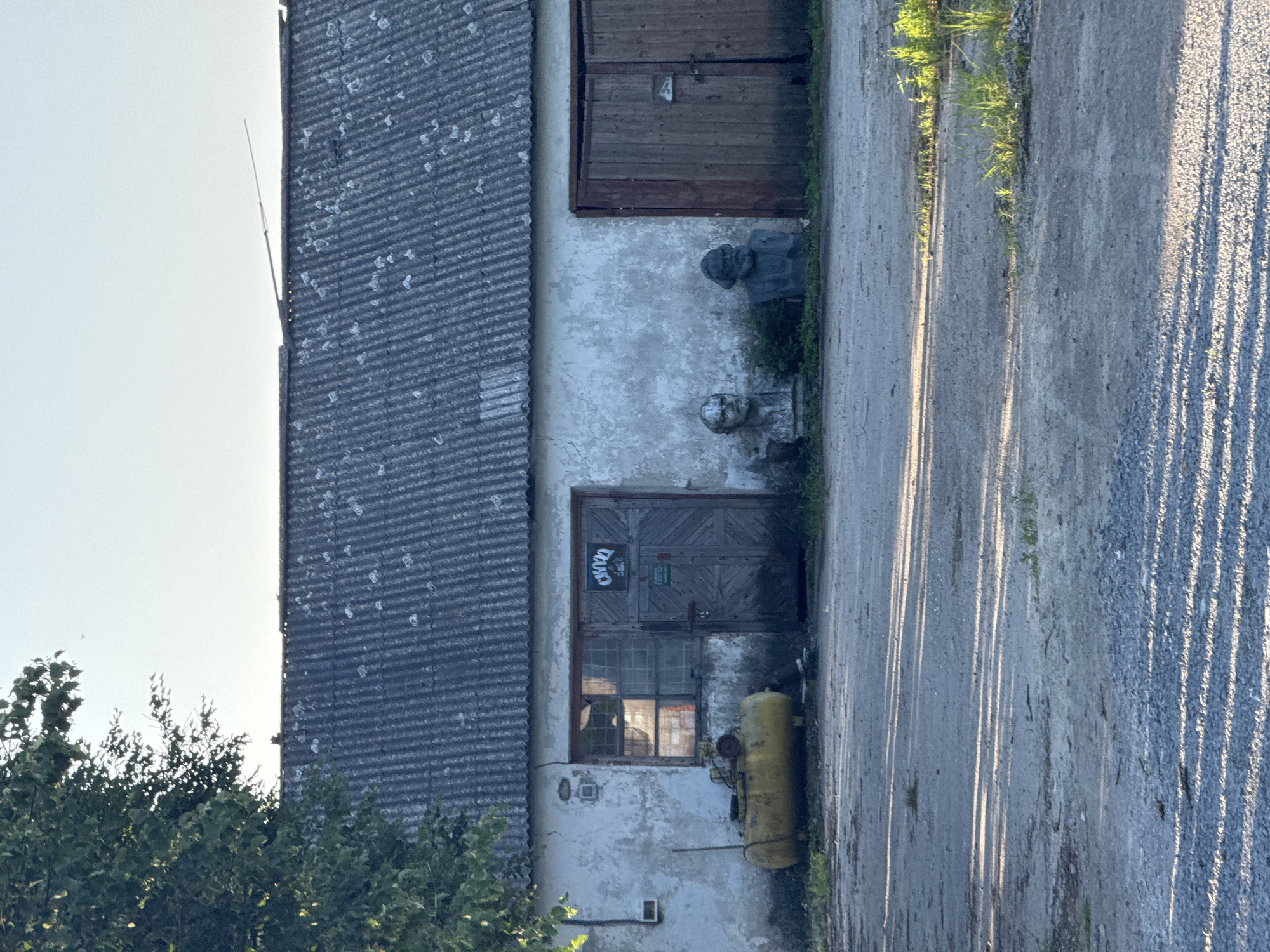 The image shows the exterior of a weathered, light-colored building with a corrugated metal roof. Two large, weathered wooden doors are visible on the right side of the building, and to their left is a smaller set of double wooden doors next to a window with multiple panes. In front of the building, to the left of the double doors, sits a large, horizontal yellow air compressor. Two busts, one of Lenin and another resembling Karl Marx, are placed on the ground in front of the building between the window and the right set of doors. Greenery and weeds grow around the bases of the busts and along the front of the building. The ground in front of the building is a gravel and dirt surface, with long shadows cast across it from an unseen light source. A large tree with dark green foliage is partially visible on the left side of the image, overhanging the building. The scene appears to be an outdoor yard or industrial area.