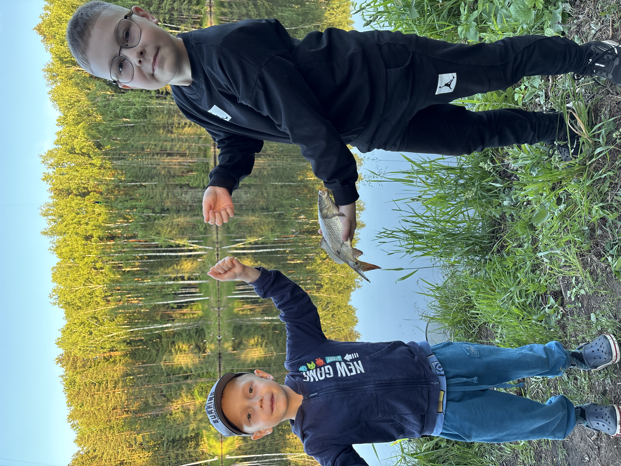 The image depicts two males, identified as children, standing on a grassy bank beside a calm body of water, likely a lake or pond, in Мишкино, Russia. The background features a dense forest composed of both coniferous and deciduous trees, with their reflections visible on the water's surface under a clear, light blue sky. The lighting suggests late afternoon or early evening.
On the left, a younger male child, estimated to be between 4 and 6 years old, faces forward. He wears a grey baseball cap with "SURFING" embroidered on the front, a dark blue zip-up hoodie featuring a multicolor graphic depicting "NEW GAME" and pixelated figures, blue sweatpants, and dark blue Crocs worn over patterned socks. His right arm is raised with his fist clenched, and a thin, barely visible fishing line extends upward from his hand. His facial expression indicates a slight smile.
On the right, an older male child, estimated to be between 8 and 12 years old, stands facing forward, tilted slightly to his left. He wears dark-framed eyeglasses, a black long-sleeved sweatshirt with a small white square logo on the upper left chest, black sweatpants with a white square Air Jordan logo on the right thigh, and black sneakers. He holds a silver-grey freshwater fish, approximately 20-25 cm long, horizontally in his cupped hands, presenting it towards the viewer. His left hand holds a fishing line, from which a green and orange fishing bobber is visible. His facial expression also indicates a slight smile.
A fishing net is partially visible on the grass behind the younger boy, near the water's edge. The ground beneath the boys is uneven and covered with green grass and various plants. Both children are looking directly at the camera.
