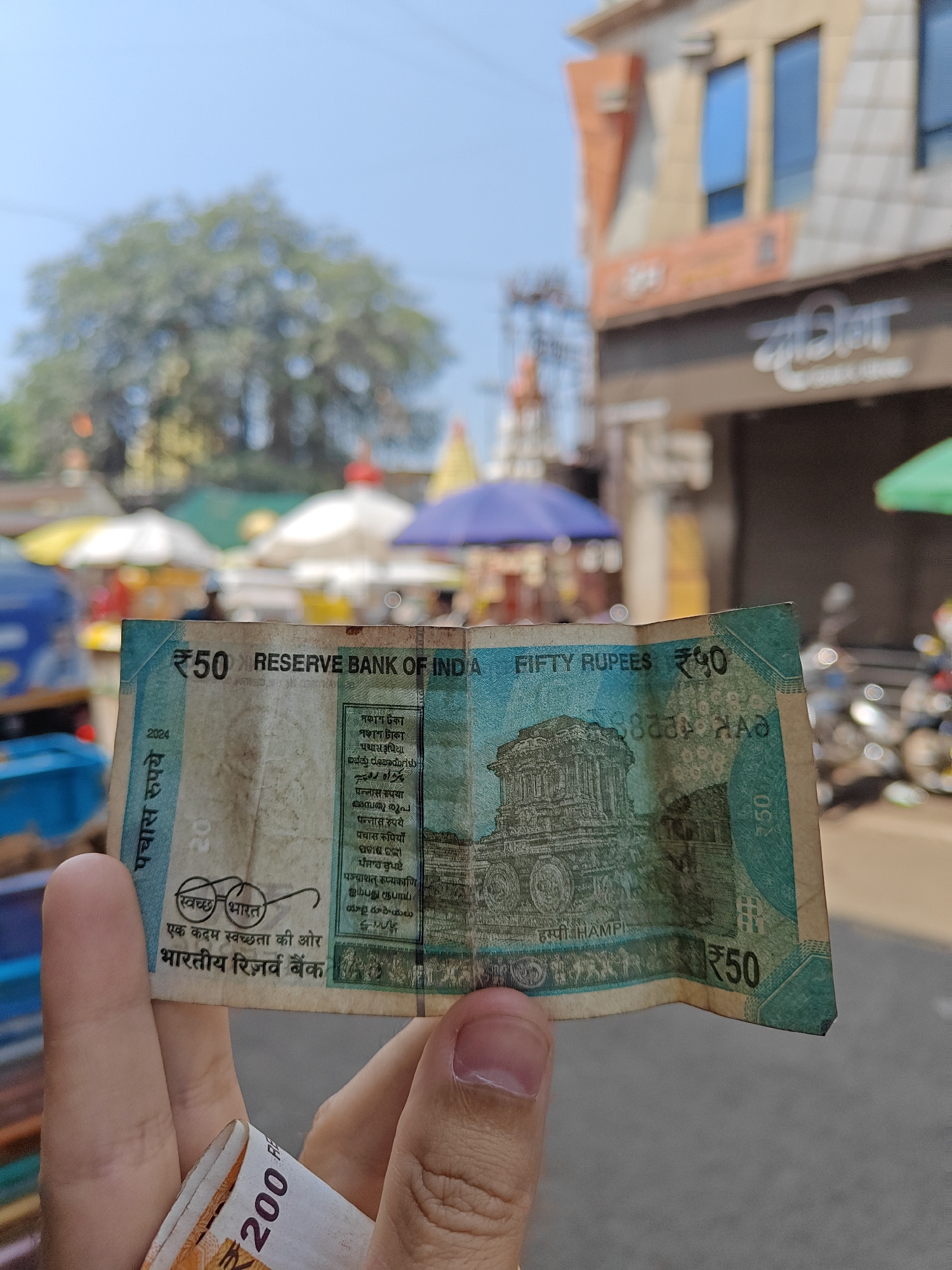 A human hand, centrally positioned, holds two Indian Rupee banknotes in an outdoor, sunlit street environment in Unknown, Unknown. The prominent banknote is a folded and creased blue-green ₹50 note, displaying "RESERVE BANK OF INDIA," "₹50" denominations, and an image of the Stone Chariot from Hampi. Beneath it, partially rolled, is an orange-colored ₹200 note.
The background, out of focus, reveals a bustling street or market scene. To the left, a large, leafy green tree is visible, with multiple market stalls featuring white, yellow, and blue umbrellas beneath it. Indistinct human figures are present among the stalls. To the right, a multi-story building with a light-colored facade and reflective blue windows rises. A dark brown shopfront, displaying white non-Latin script, is visible at ground level, with a motorcycle parked nearby. The sky above is clear blue.