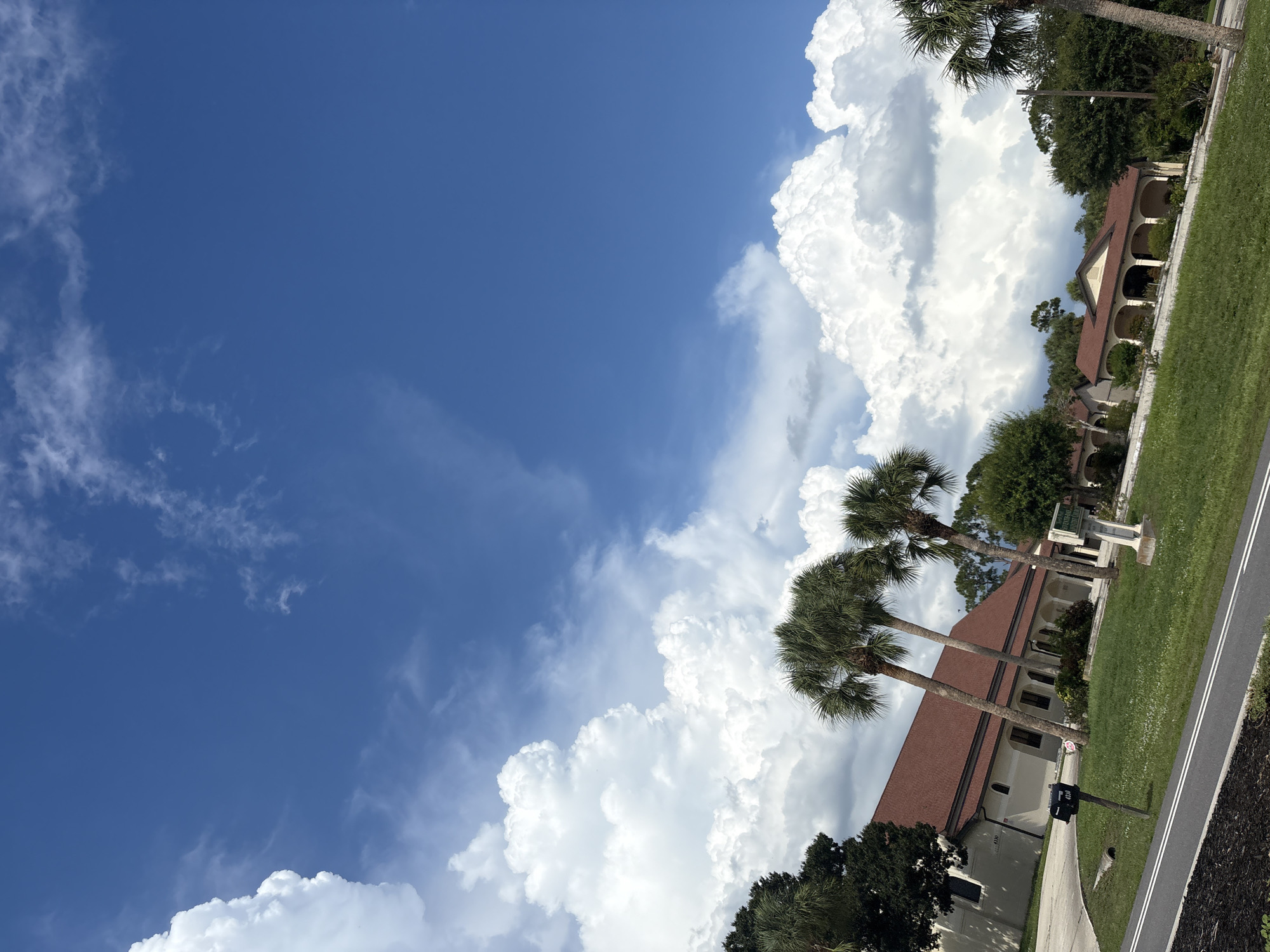 An outdoor, eye-level, medium shot captures a building with a red-tiled roof and white walls, situated in North Port, United States of America. The building appears to be a church or a similar institutional structure, with arched openings visible along its facade. Several palm trees are scattered throughout the scene, some towering over the building, while others are shorter. The sky is a vibrant blue, punctuated by large, fluffy white clouds, suggesting fair weather. In the foreground, a grassy area is visible, along with a paved pathway or road. A black mailbox with the address "4330" is positioned near the building. The overall impression is of a tranquil day in a suburban or semi-rural setting.
