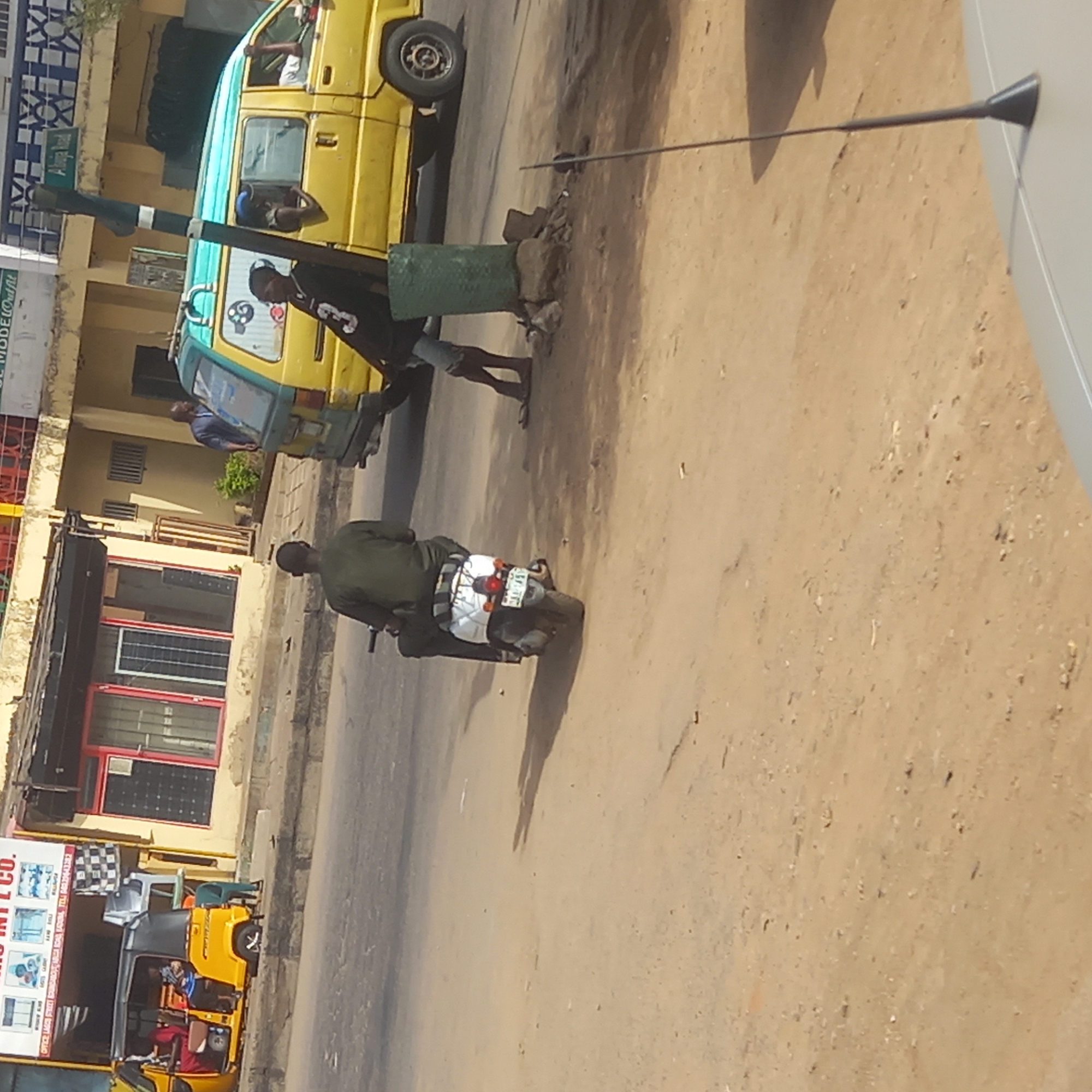 The photograph depicts a street scene in what is identified as Dadi Riba, Nigeria. A yellow and light blue passenger van is parked on the left side of the frame, with at least two individuals visible inside or near the vehicle. One person is standing on the street, leaning on a green-painted, pole-like object. A motorcyclist is riding a motorcycle with a white seat and visible license plate away from the camera. A yellow auto rickshaw (tuk-tuk) is parked in front of a business with signage. The street surface is a mix of pavement and dirt, and there's a satellite dish in the upper right corner. The overall setting appears to be a commercial or residential area within the city.